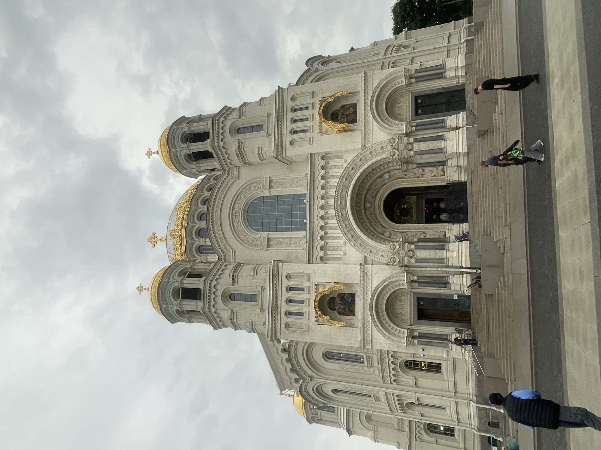 The image shows a wide-angle, low-angle shot of the Kronstadt Naval Cathedral in Kronstadt, Russia. The cathedral is a large, white building with multiple golden domes topped with crosses. The facade features intricate carvings, arched doorways, and tall windows. Several people are visible on the steps leading up to the cathedral and walking on the plaza in front of it. The sky is overcast.