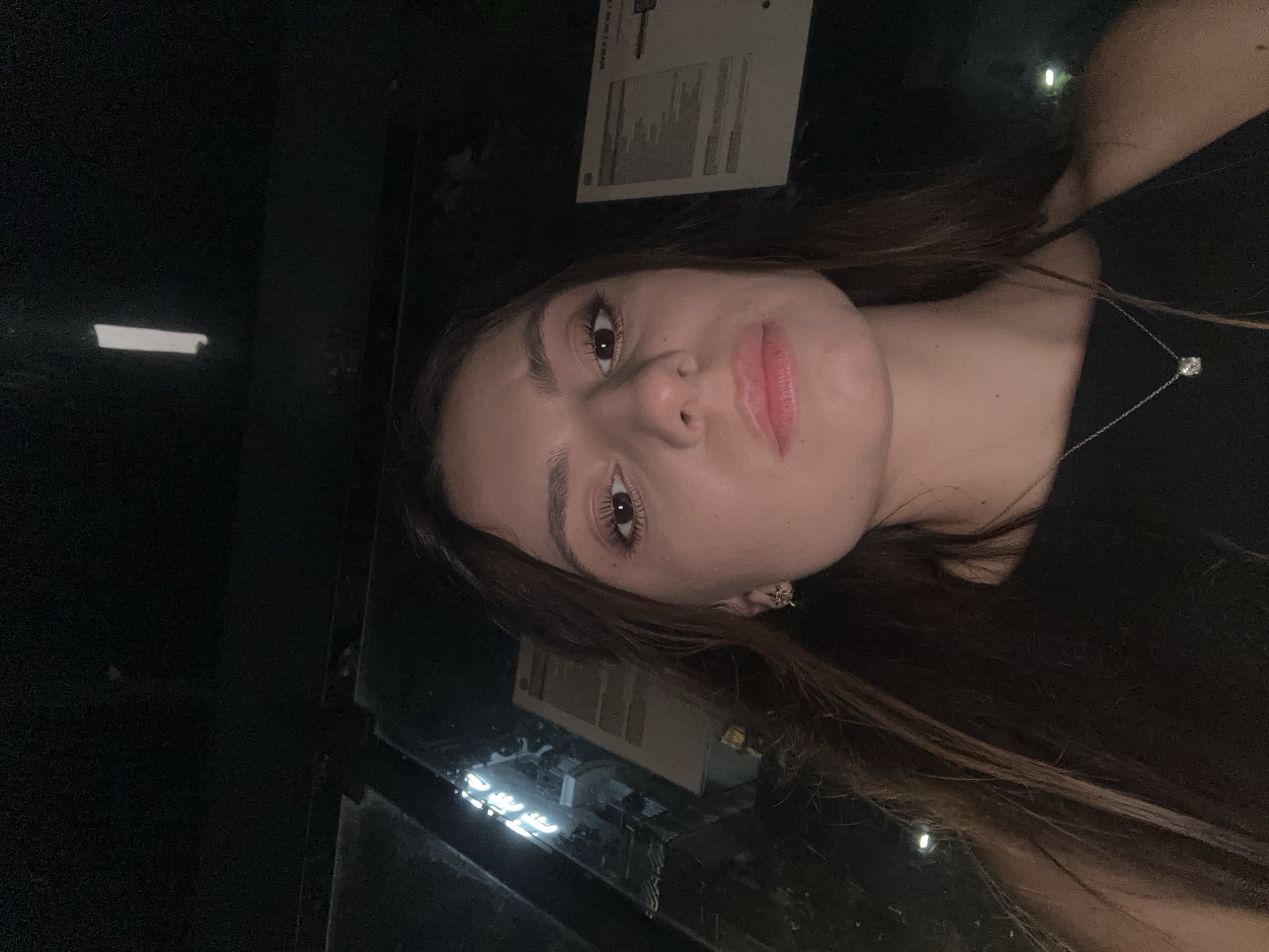 A young woman with long, dark brown hair is taking a selfie in what appears to be a dark interior. She is wearing a black top and a delicate silver necklace with a single clear pendant. Her expression is neutral. Behind her, the background is dark and indistinct, with some faint reflections or lights visible. The overall lighting is dim, suggesting the photo was taken at night or in a dimly lit environment. It is not possible to determine the specific location in Varna, Bulgaria from the image provided.