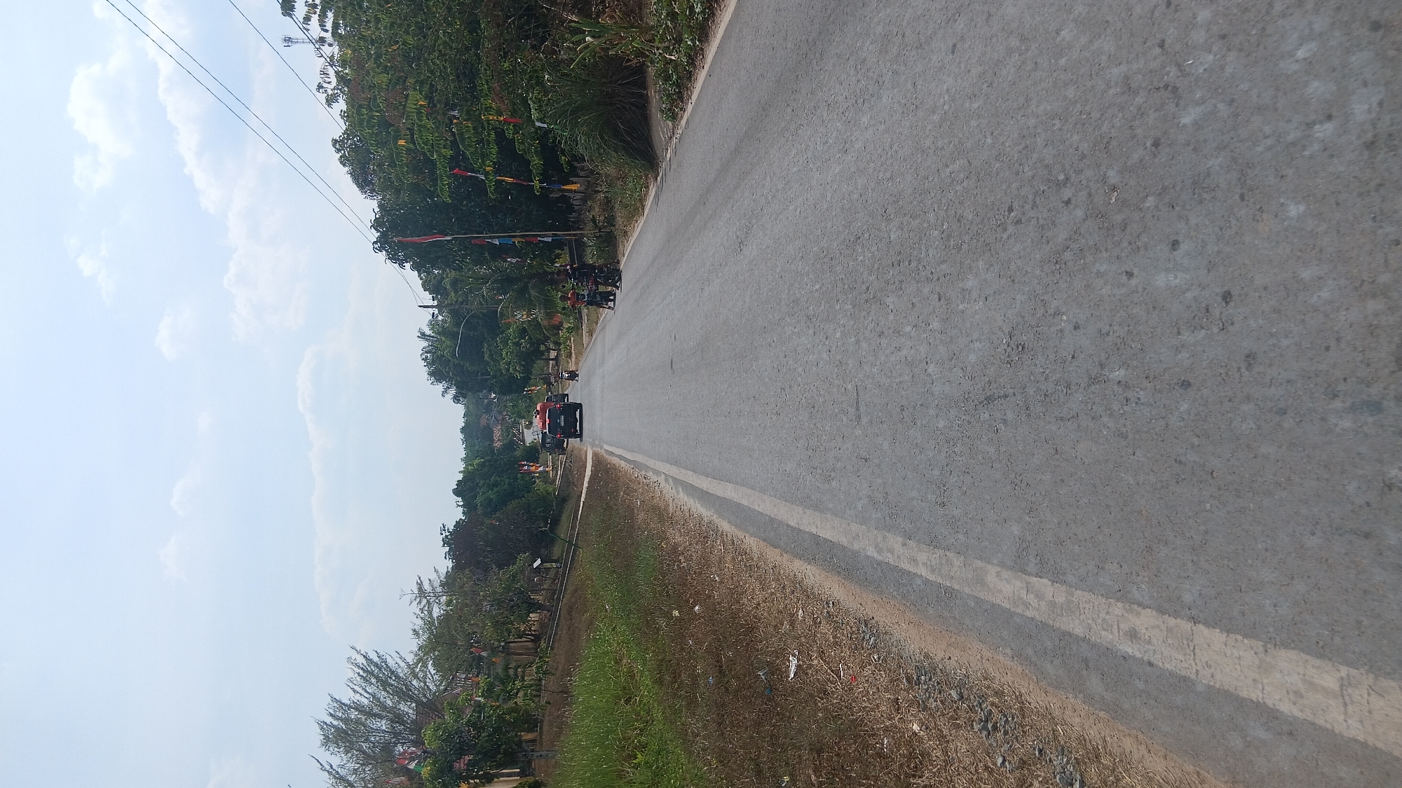 The image depicts a two-lane asphalt road in Prabumulih, Indonesia. The road is bordered by dry grass and vegetation on the left and trees and houses on the right. Several vehicles are visible on the road, including a red truck, a black SUV, and two motorcycles with riders. A few people are standing on the side of the road near the motorcycles. Colorful flags are strung along the sides of the road, suggesting a festive or celebratory occasion. The sky is partly cloudy and the overall lighting suggests it is daytime.