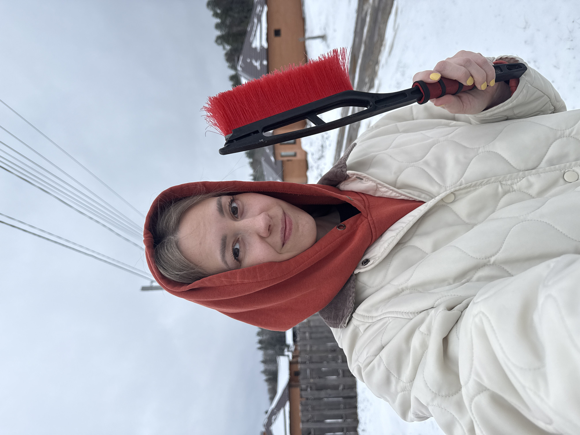 A young woman in a red hoodie and a white quilted jacket holds a red and black snow brush. The woman is smiling at the camera. It is daytime and the sky is overcast. Powerlines can be seen in the upper left corner. There is snow on the ground. In the background, there are trees and buildings, possibly wooden structures. Based on the location, this image was likely taken in a snowy, suburban setting near Chaikovsky, Russia.