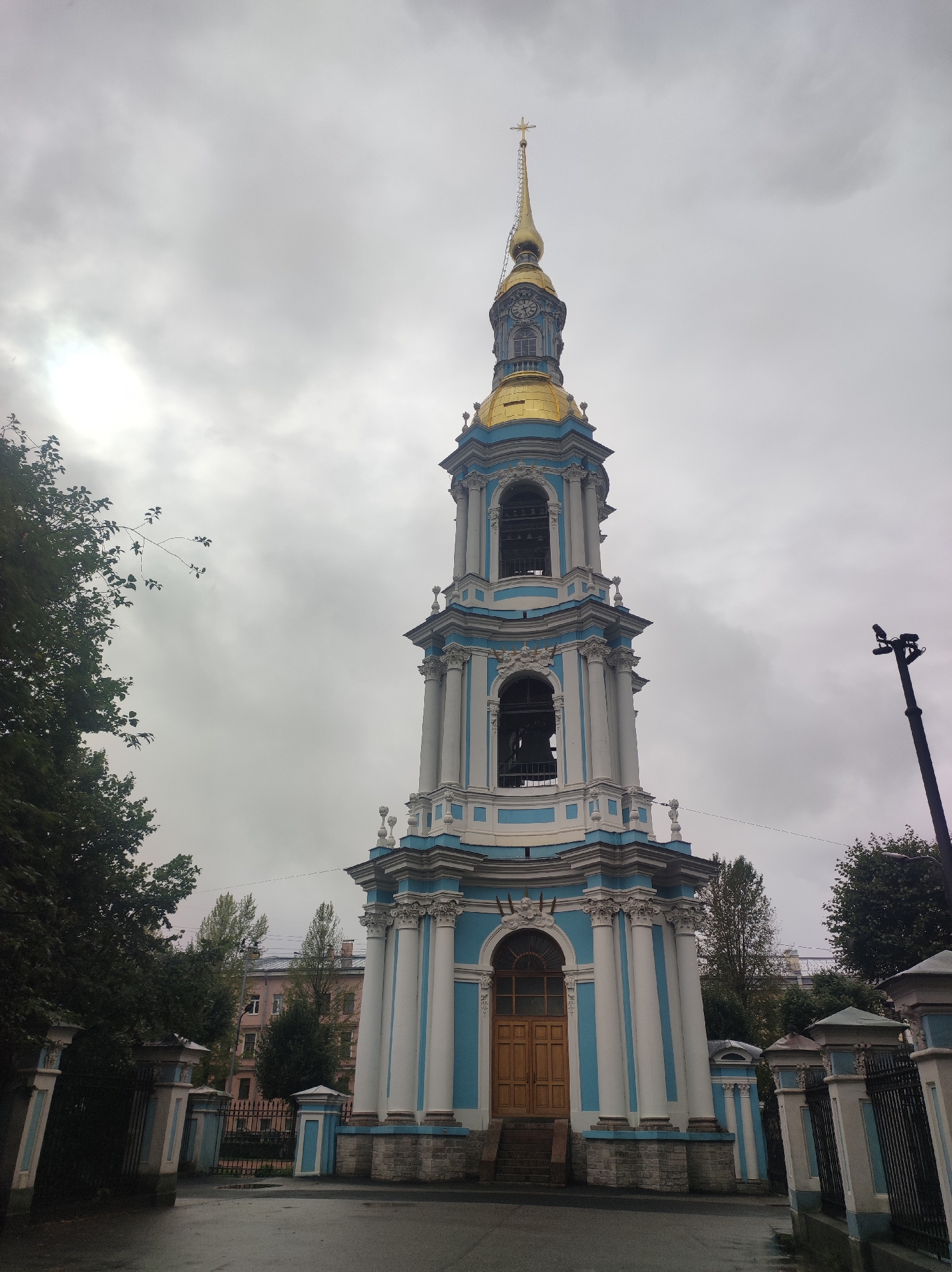 This is a full-shot, eye-level, outdoor, natural-light photograph of the bell tower of Saint Nicholas Naval Cathedral in Saint Petersburg, Russia, on a cloudy day. The bell tower is a tall, multi-tiered structure painted white with blue accents and topped with a golden spire and cross. The tower features multiple arched openings, some of which contain bells, and is decorated with columns and intricate moldings. A large wooden door is visible at the base of the tower, accessible by a set of stairs. The tower is surrounded by a fenced courtyard with decorative pillars. To the left of the tower, lush green trees partially obscure the view, while to the right, a tall lamppost with a security camera is visible. In the background, residential buildings and more trees can be seen. The ground in front of the tower appears wet, suggesting recent rain. There are no people visible in the image.
