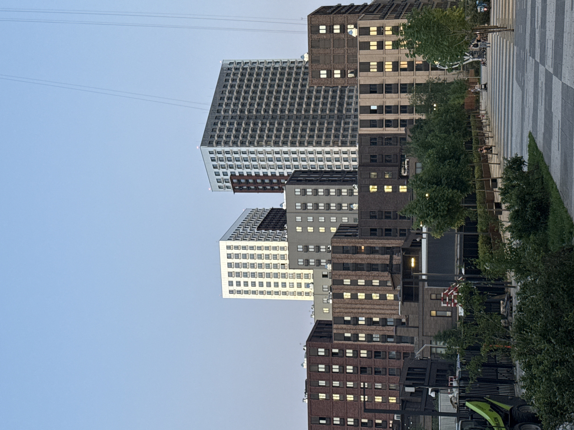 The image shows a cityscape, likely in Moscow, Russia, featuring a cluster of buildings with distinct architectural styles. Several buildings have a uniform window grid design with reflective surfaces. Some buildings have a more traditional brick facade with rows of rectangular windows, some of which are illuminated. Power lines stretch across the top of the frame. The buildings are seen from a street level. Trees and some bushes are visible in the foreground. There is a sidewalk with a checkered pattern to the right. People are sitting on benches. In the bottom right of the image, there appears to be some construction equipment.