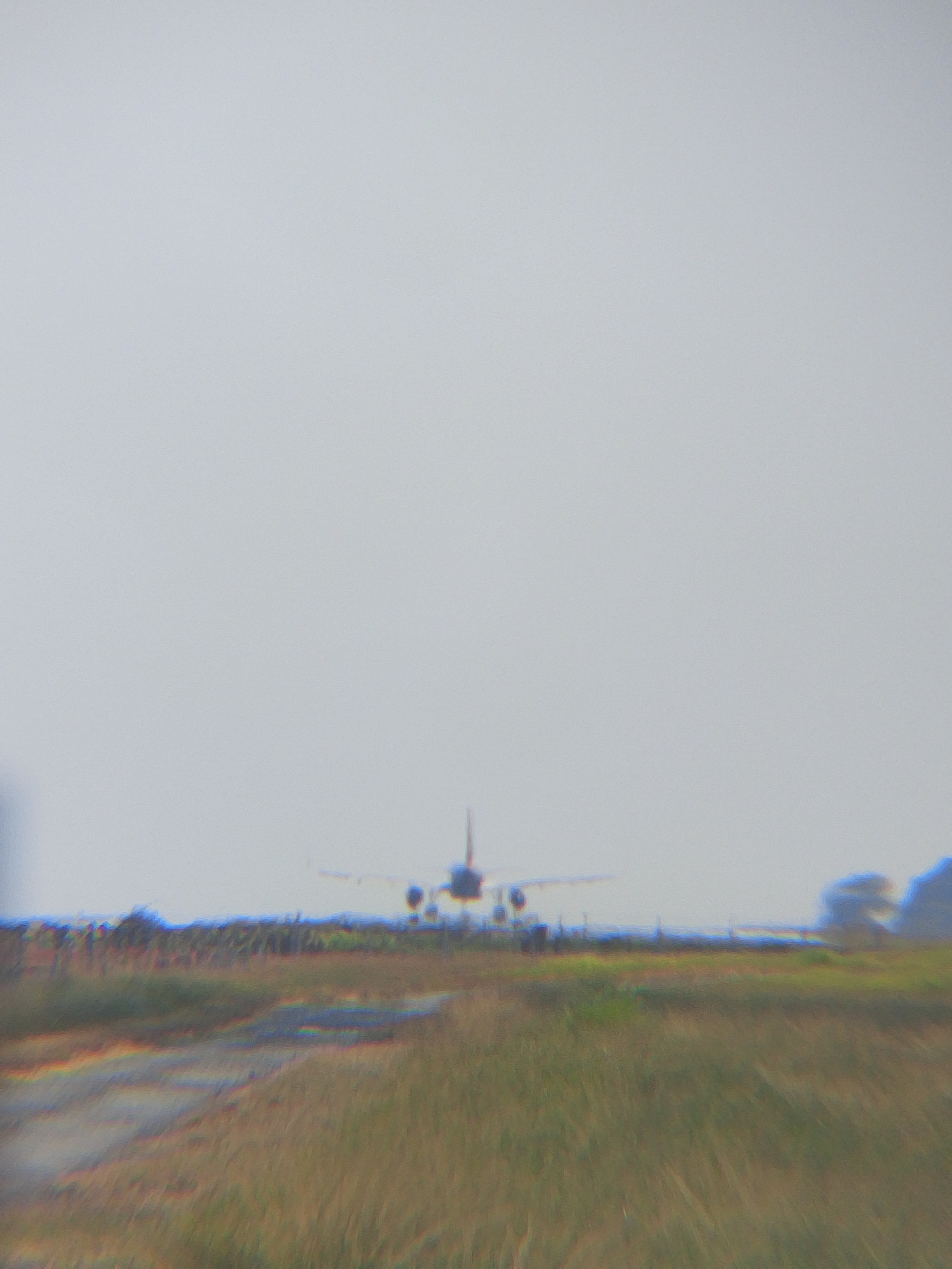 The image captures an airplane in mid-air against a hazy sky, appearing to be either taking off or landing. The foreground consists of a grassy field with a dirt path or road running through it. The lower portion of the image shows blurry streaks of color, possibly indicative of motion blur or atmospheric conditions. The airplane is the primary subject, silhouetted against the lighter sky. The scene appears to be an outdoor environment, possibly near an airport or an open field. Without specific landmarks or identifying features, it is not possible to determine the exact location within Balikpapan, Indonesia.