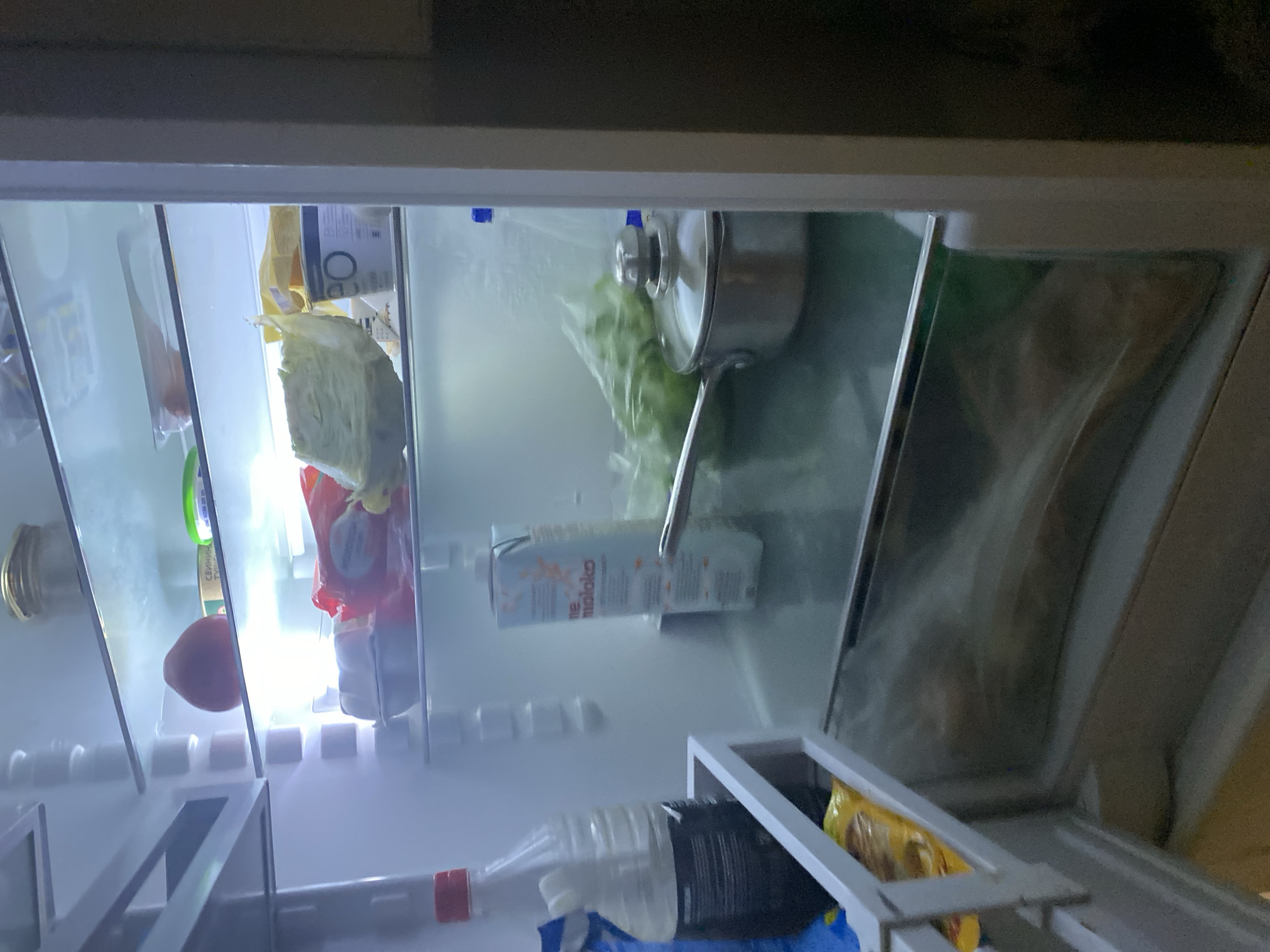 The image shows the interior of a refrigerator, illuminated by interior lights. The refrigerator contains various food items. On the top shelf, there is a glass jar and a red, round fruit. Below that, on the next shelf, are packages of what appear to be food items, including a white container and a red and white package. Further down, a large piece of lettuce is visible. On a lower shelf, there is a carton of what appears to be milk, and a stainless steel pot with a lid. To the left, a plastic bottle with a dark liquid is present. The bottom of the refrigerator contains some items in plastic bags. The image is a static interior shot, with no people or discernible actions. The location of Tolyatti, Russia, is a context provided and not directly inferable from the visual content of the image itself.