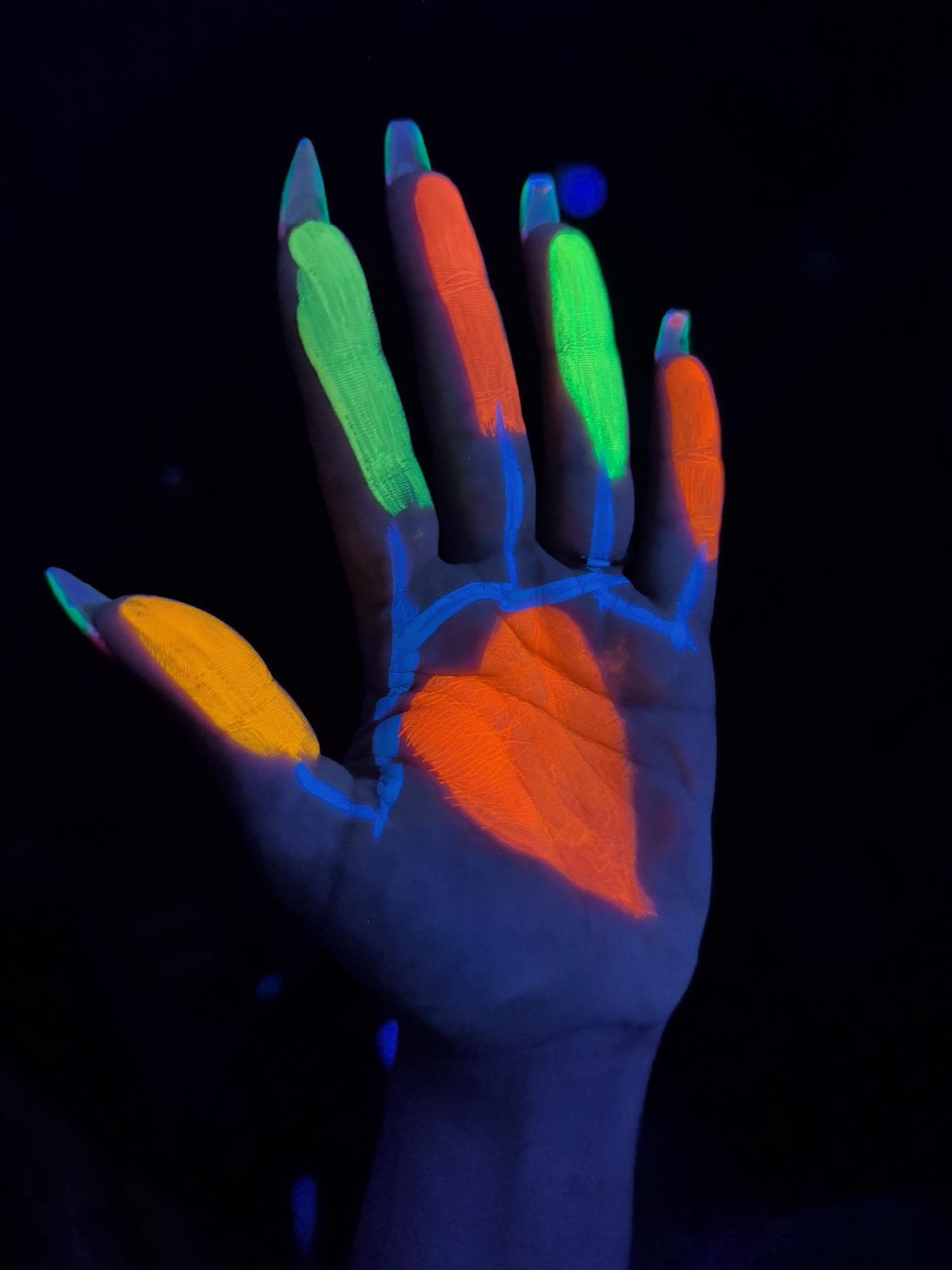 A hand is presented under UV light. The palm is colored orange. The thumb is colored orange. The index and ring fingers are colored green. The middle and pinky fingers are colored orange. The fingers, palm and wrist are connected by blue lines that appear to be veins. The background is black. The photo does not contain information about the location.