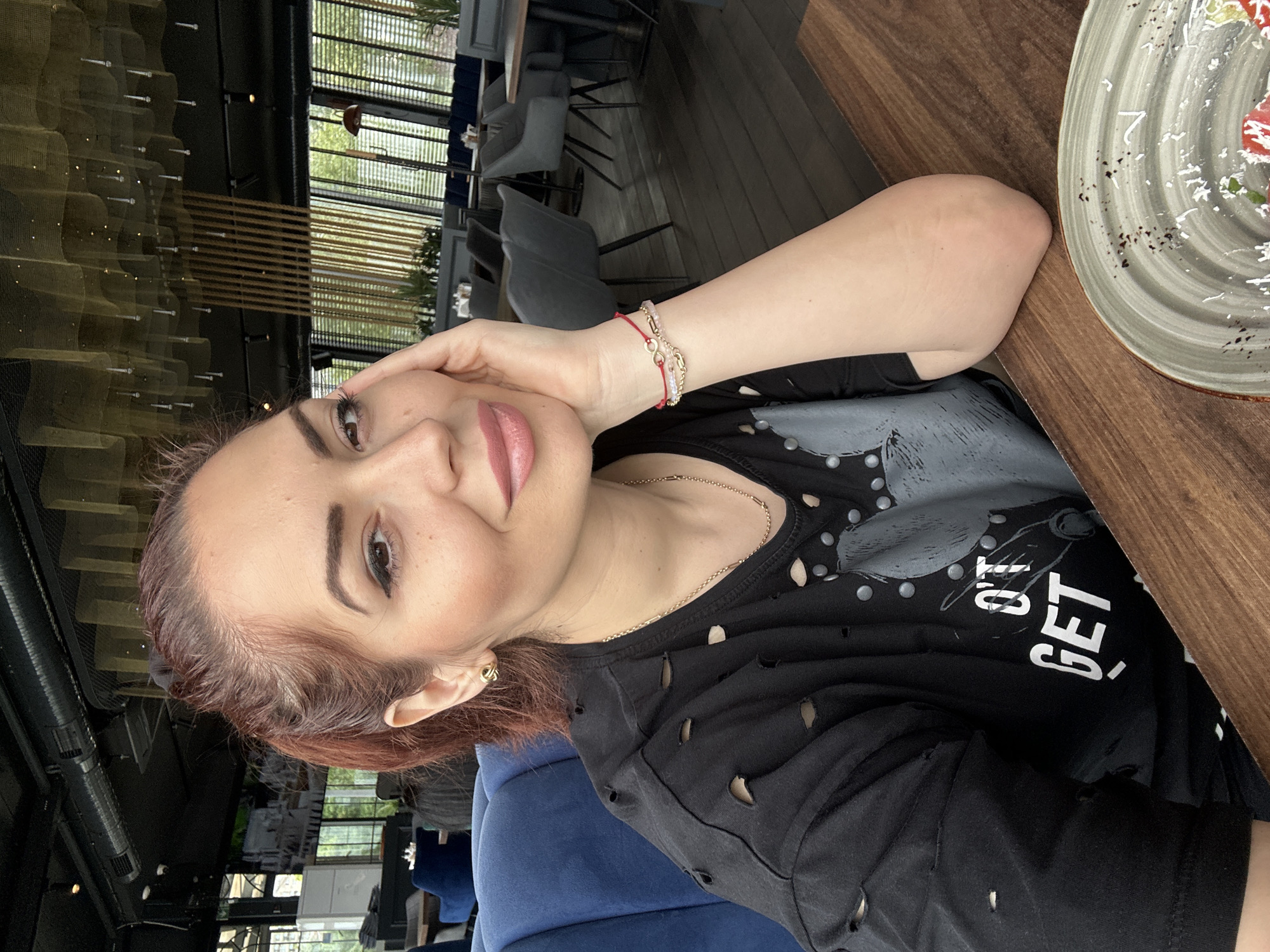 A woman is seated at a wooden table in what appears to be a restaurant. The interior is modern, with black metal framing and hanging light fixtures visible in the background. Tall windows reveal greenery outside, suggesting an outdoor or garden setting. The woman is wearing a black distressed shirt with a print that includes the word "GET" in white letters. She has a gold chain necklace and a bracelet with red and gold elements on her wrist. A plate with remnants of food is on the table beside her. The location could be a restaurant in the city of Vidin, Bulgaria based on architectural styles and typical outdoor restaurant setups.