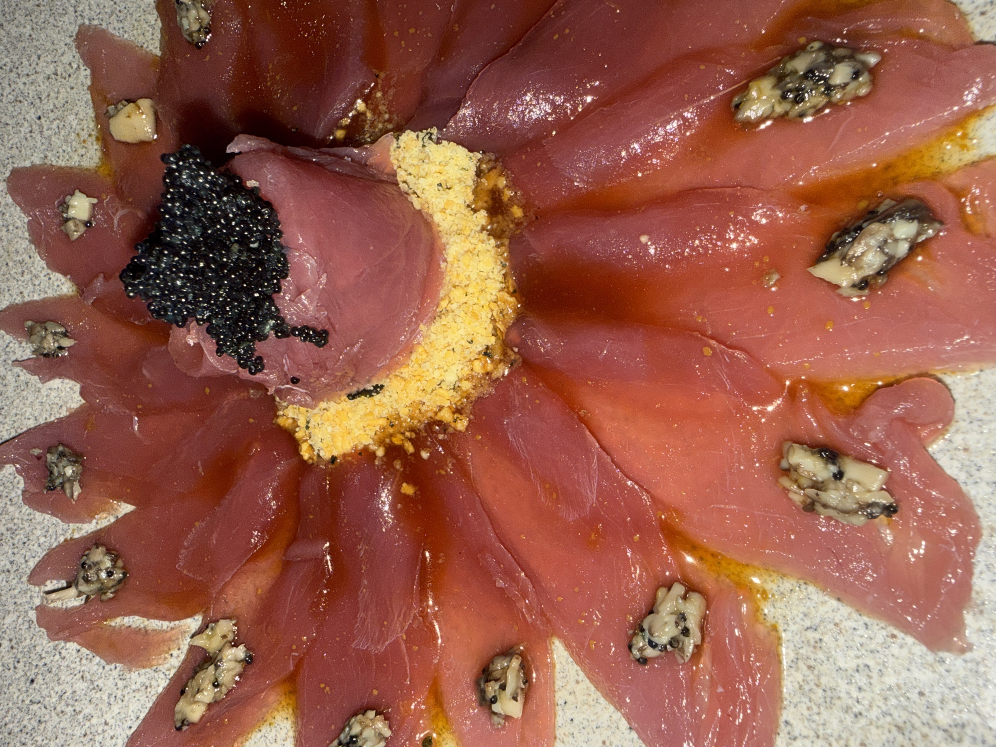A finely plated gourmet dish is presented on a light grey, speckled plate. The main subject is thin slices of reddish-pink raw fish, likely tuna, arranged in a circular, flower-petal-like pattern.
In the center of the dish, a small mound of the same raw fish is topped with a generous portion of glossy black caviar. Adjacent to this central mound, and filling the crevices between the radiating fish slices, is a granular, yellowish-orange ingredient, possibly grated bottarga or cured egg yolk.
Many of the individual fish slices are garnished with small, irregular clusters of a greyish-white, finely chopped ingredient, which contains small black specks. This could be a type of relish, chopped shellfish, or mushrooms. The entire arrangement is coated in a thin layer of glossy, reddish-brown liquid, indicative of a dressing or sauce.
No people are visible. The setting is indoors, implied by the focus on the prepared food. There is no visible text or specific location cues within the image.
