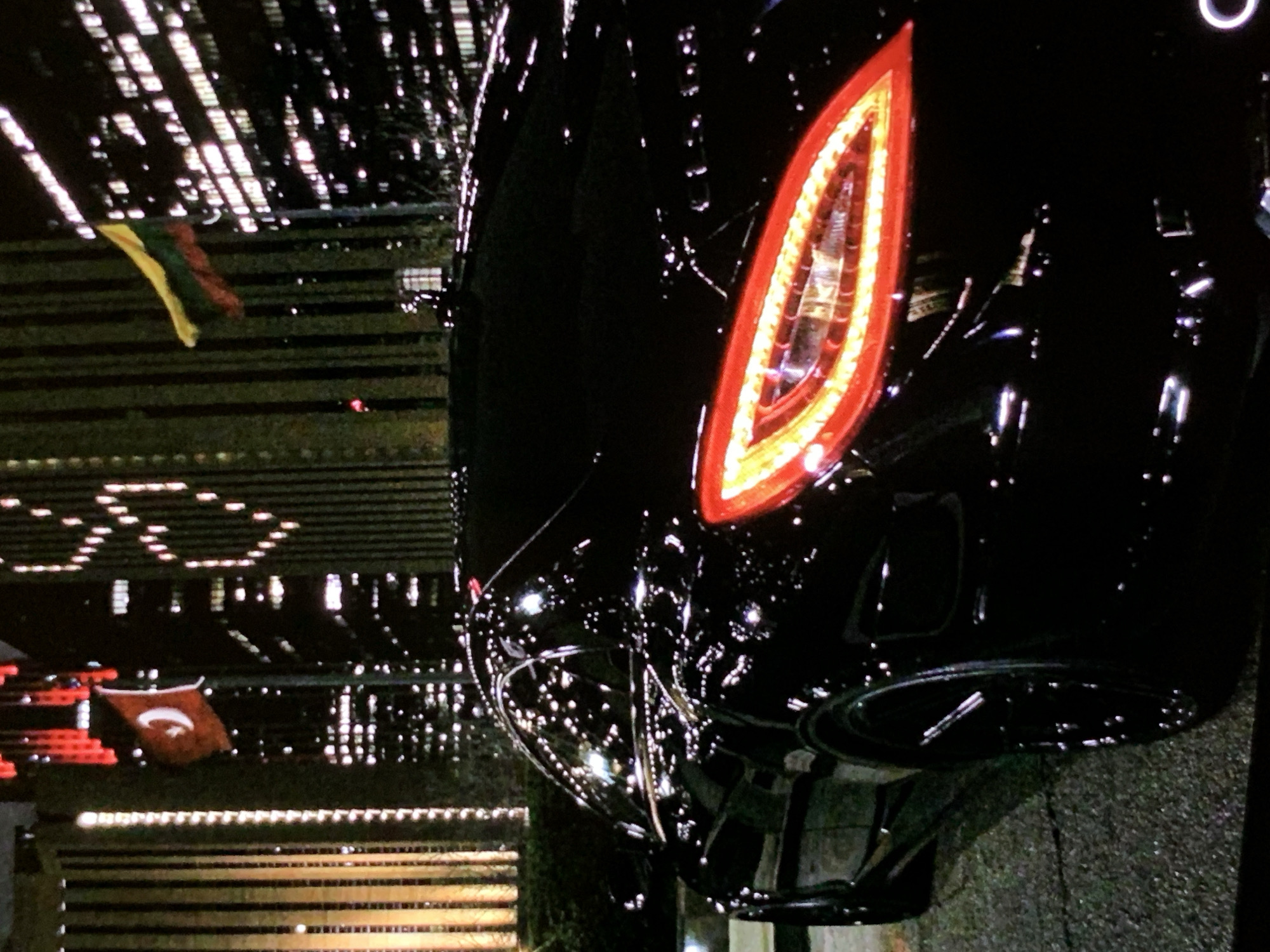 In a nighttime urban setting, a black luxury sedan is parked on a wet asphalt surface. The car's red taillights are illuminated, reflecting off its polished black exterior. In the background, tall buildings are adorned with numerous lights, some forming patterns. Two flags are visible on flagpoles: one is a red flag with a white crescent and star, and the other is a tricolor flag with yellow, green, and red stripes. The scene appears to be at street level, with what looks like a hedge to the left of the vehicle.