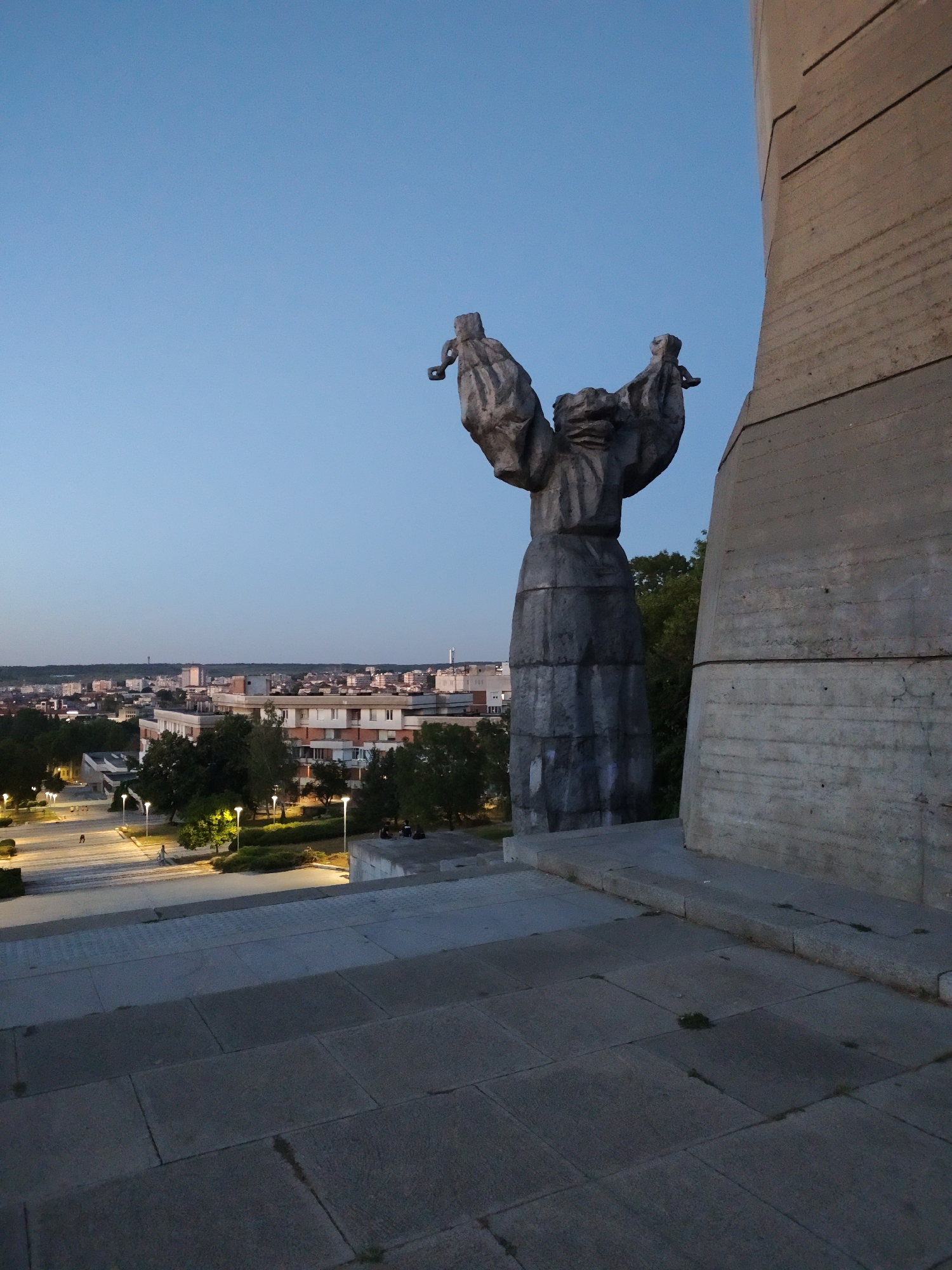 The image captures a view from a high vantage point overlooking the city of Pleven, Bulgaria at dusk. In the foreground, a large, dark gray, abstract statue stands prominently to the left of a massive concrete structure. The statue appears to be made of segmented stone or concrete, with outstretched arms holding what look like chains or ropes. The concrete structure is to the right, its rough texture and horizontal banding clearly visible.
Beyond the immediate foreground, a wide, stone-tiled plaza or walkway slopes downward. Several modern streetlights, emitting a warm yellow glow, illuminate the plaza and the surrounding areas. In the middle ground, a cityscape unfolds with a variety of buildings, mostly residential or commercial structures, with varying architectural styles and heights. Green trees are interspersed throughout the city, and the distant horizon shows a hazy outline of more buildings and possibly hills.
In the plaza, a few individuals are visible. Two people are walking up a set of stairs on the left side of the plaza. Further to the right, near a cluster of trees, a small group of people are seated. The sky above is a clear, soft blue, transitioning to a deeper hue near the horizon, indicating the time is just after sunset. The overall impression is one of a monument overlooking a city at twilight.
