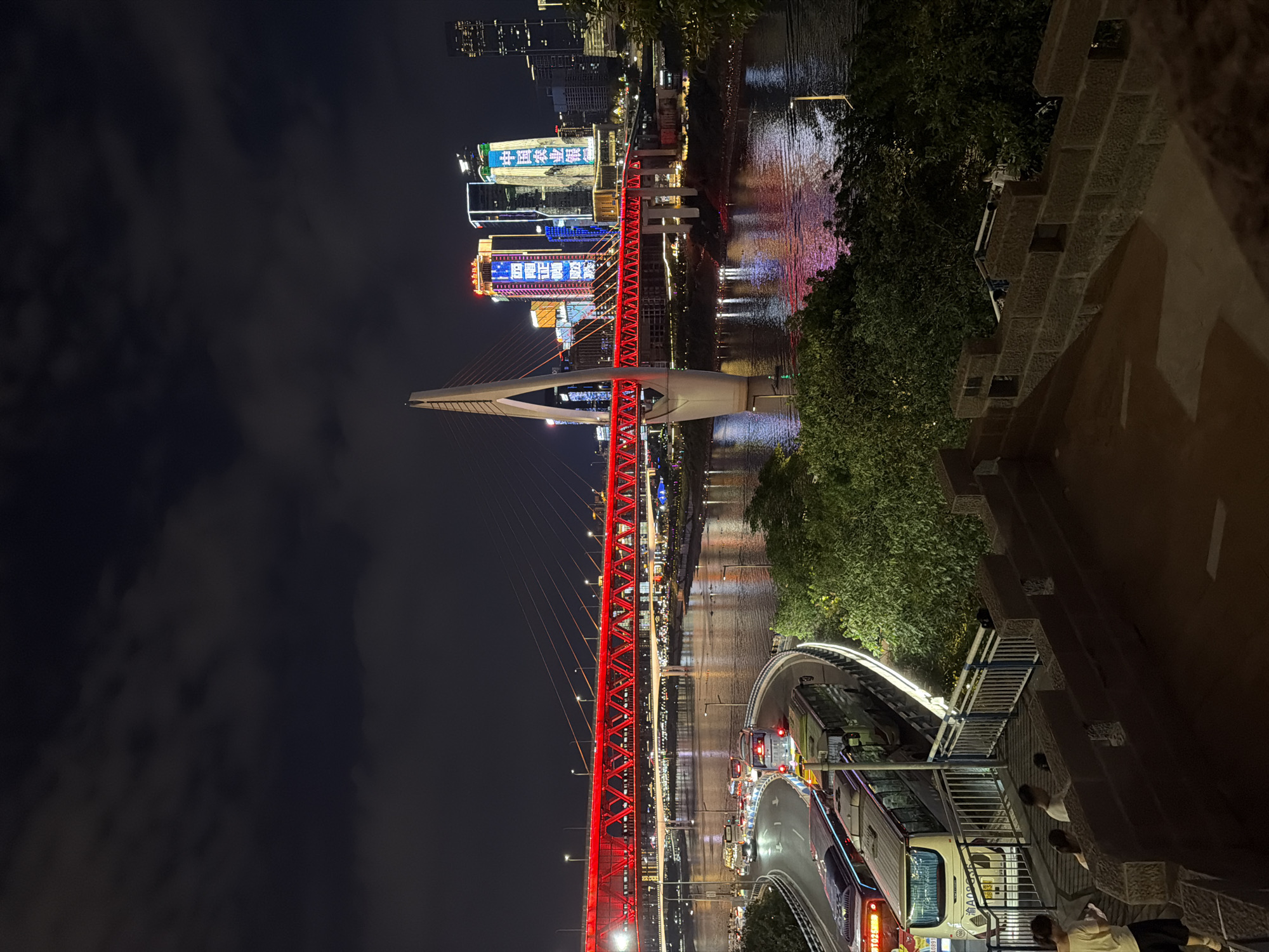 An eye-level, medium shot captures a night scene in Yuzhong District, China, featuring a river, a bridge, and city buildings. The foreground is dominated by a stone wall with square openings, with a set of stairs descending to the left. On the stairs, two women and a man are visible, seemingly looking at their phones or devices. Below the stairs, a curved road with traffic is illuminated, with several buses lined up. The middle ground shows the river, reflecting the colorful lights of the city. A prominent suspension bridge with a large, white pylon is lit in red, spanning across the water. In the background, modern skyscrapers are adorned with bright, colorful lights and advertisements. The sky above is dark with scattered clouds, and the overall atmosphere is illuminated by artificial lights.