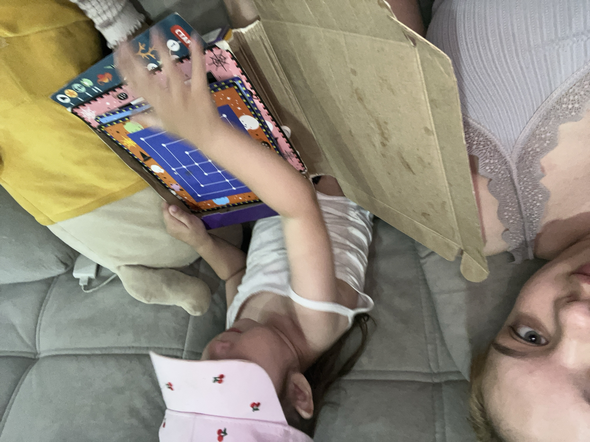 This media file shows a close-up, overhead view of a group of people on a couch. The subjects include a child lying on their back, wearing a white tank top and a pink hat adorned with cherries. Their face is partially obscured, but their eyes are visible. To the left of the child, a person wearing a yellow shirt is partially visible, along with a plush toy. To the right of the child, another person is lying on their back, facing the camera. They are wearing a light blue ribbed top with lace trim. A cardboard box is positioned between the child and the person on the right. The child is holding a colorful board game, possibly a puzzle, with a blue grid pattern. The image appears to be taken indoors, likely in a home setting, given the couch and the presence of a plush toy. The lighting suggests it could be daytime. The overall mood seems relaxed and casual, possibly during playtime or a quiet moment at home in Gorodets, Russia. The board game has some visible text, including "CTA" and numbers.