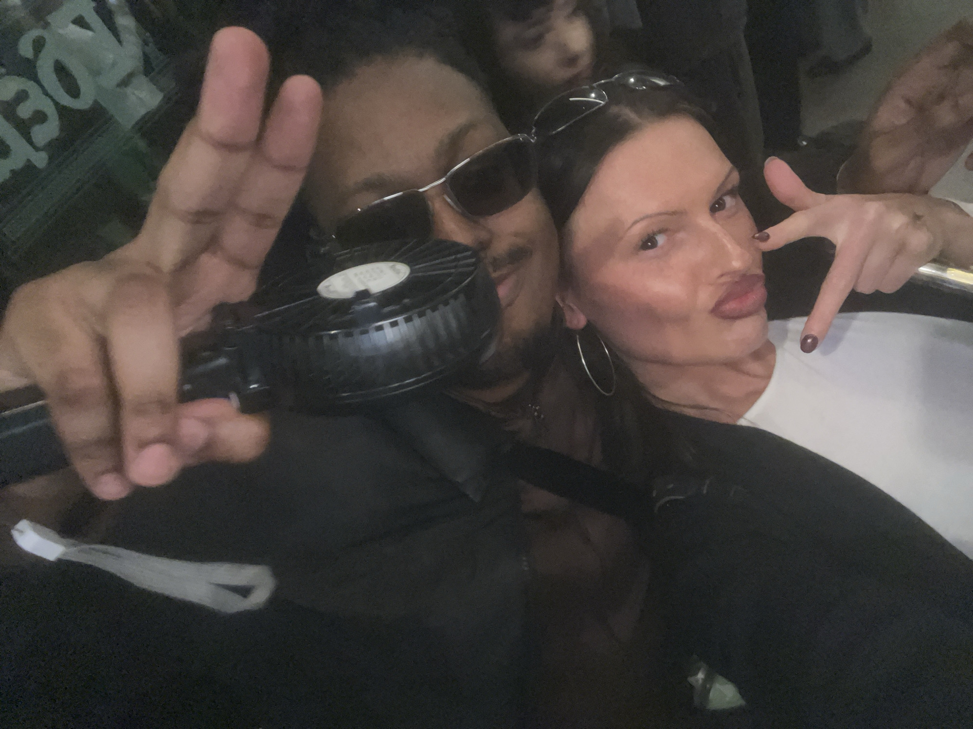 A dim, close-up selfie captures two adults in the foreground, with a third person partially visible behind them. On the left, a man with a dark complexion, wearing sunglasses, raises his left hand in a peace or victory sign. He holds a black, circular device with a grid pattern, resembling a portable fan or speaker, from which a light-colored strap hangs. Faint, reversed green text, appearing as "3JDe", is visible in the background over his left shoulder. To the right, a woman with dark hair, wearing sunglasses pushed up onto her head and large hoop earrings, makes a "duck face" towards the camera. She extends her right hand, pointing downwards with her index finger while her thumb is up; her fingernails are painted dark. The setting appears to be an indoor, dimly lit environment, suggesting a social venue or nightlife activity. The poses convey a casual and playful mood.