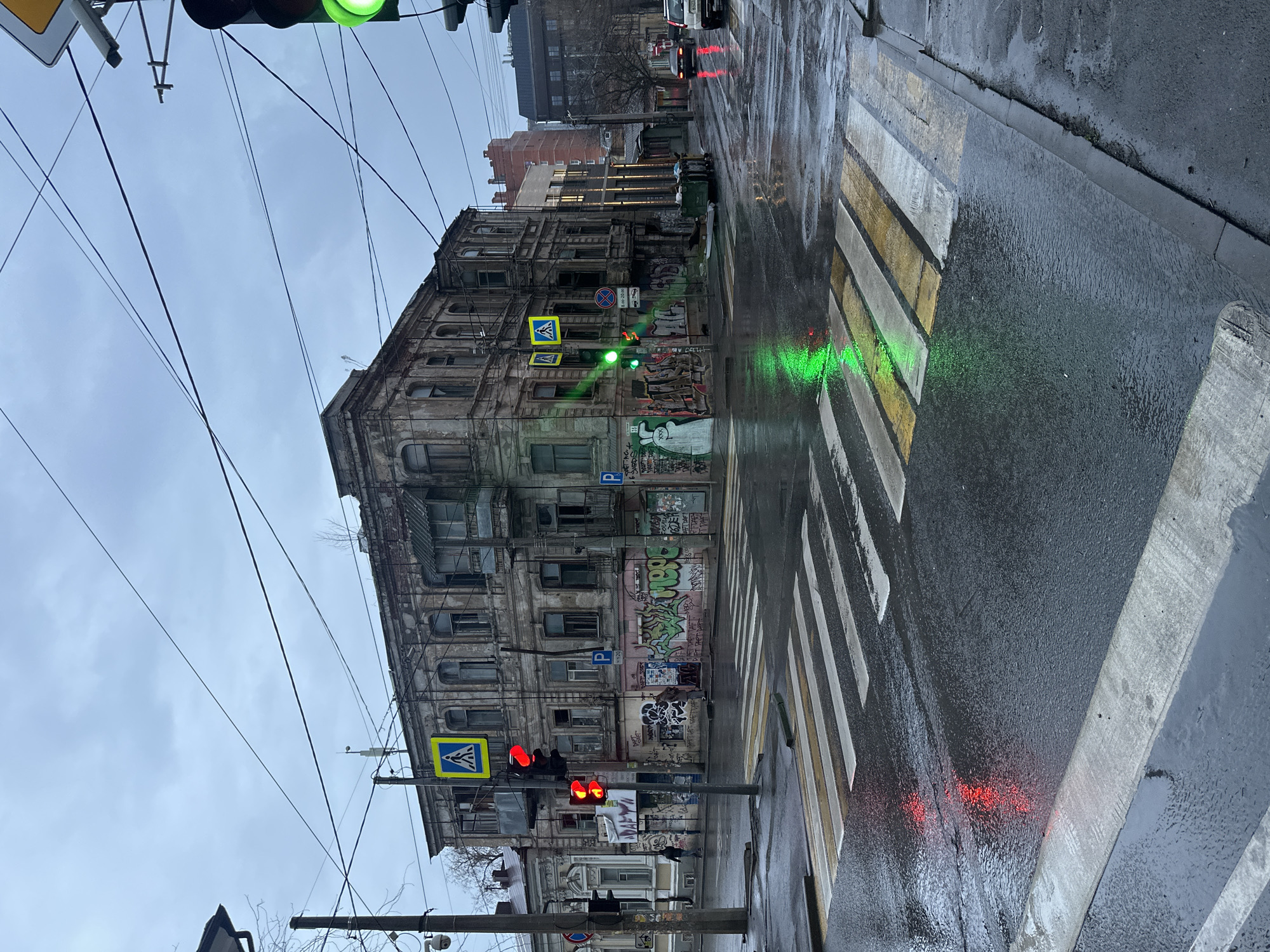 The image depicts a street scene in Rostov-on-Don, Russia, on a wet, overcast day. The focus is on an old, dilapidated building covered in graffiti, situated at a crosswalk. Traffic lights indicate red in one direction and green in the other, reflecting off the wet pavement. A pedestrian crossing sign is visible. There are power lines overhead. The overall environment appears urban and somewhat neglected.