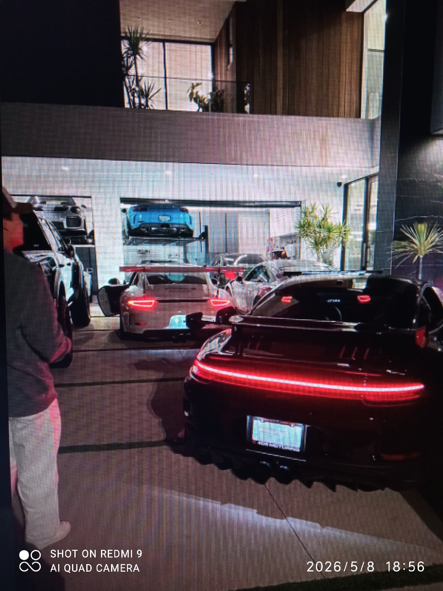 The image depicts a residential scene at night in an unknown city. The focus is on several high-end vehicles parked in front of a modern, multi-story house with large windows and potted palm trees. In the foreground, the rear of a black Porsche with illuminated tail lights is visible, with its license plate partially obscured. Behind this, a white Porsche with its door open is parked. A blue car is seen elevated on a car lift within the garage. A person, with their back to the camera, stands to the left of the frame. The ground is paved with rectangular slabs. The date "2026/5/8 18:56" is stamped on the image. A "SHOT ON REDMI 9 AI QUAD CAMERA" watermark is also present.