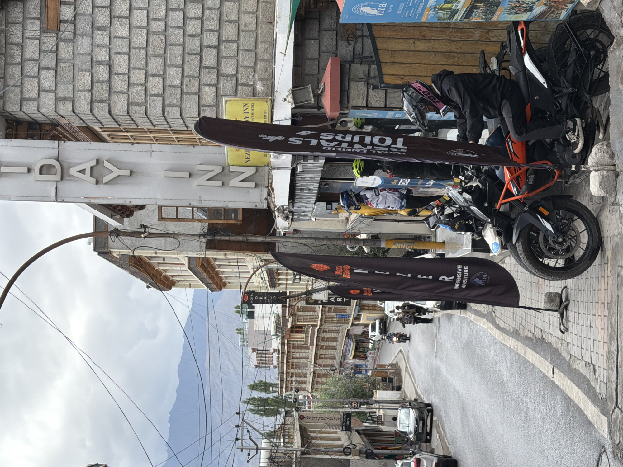 The image captures a street scene in the city of Leh, India, under a cloudy sky. In the foreground, a person wearing a black jacket, helmet, and track pants is seated on an orange and black motorcycle. The motorcycle has a black flag with white text that reads "ROYAL ENFIELD TRIALS TOURS" and "INEXPENSIVE ADVENTURE" positioned to its left. Another person is visible behind the first, partially obscured.
To the right of the motorcycle, a building with a grey brick facade is prominent, featuring a sign that reads "HIDDEN INN" vertically. Next to this sign, a smaller sign states "NEZELAY INN (GUEST HOUSE)". Further to the right, a blue poster is affixed to a wooden structure, displaying text and images related to the region, including "MA SINCE 1985" and "ORGANIZED LADAKH".
The street itself is paved and curves uphill, with several buildings lining both sides. Cars and motorcycles are present on the road, with a few people walking. In the background, distant mountains are visible, their slopes muted by the overcast conditions. The environment suggests a town or city setting with a blend of commercial establishments and residential buildings. Power lines are strung across the sky, a common sight in urban areas.