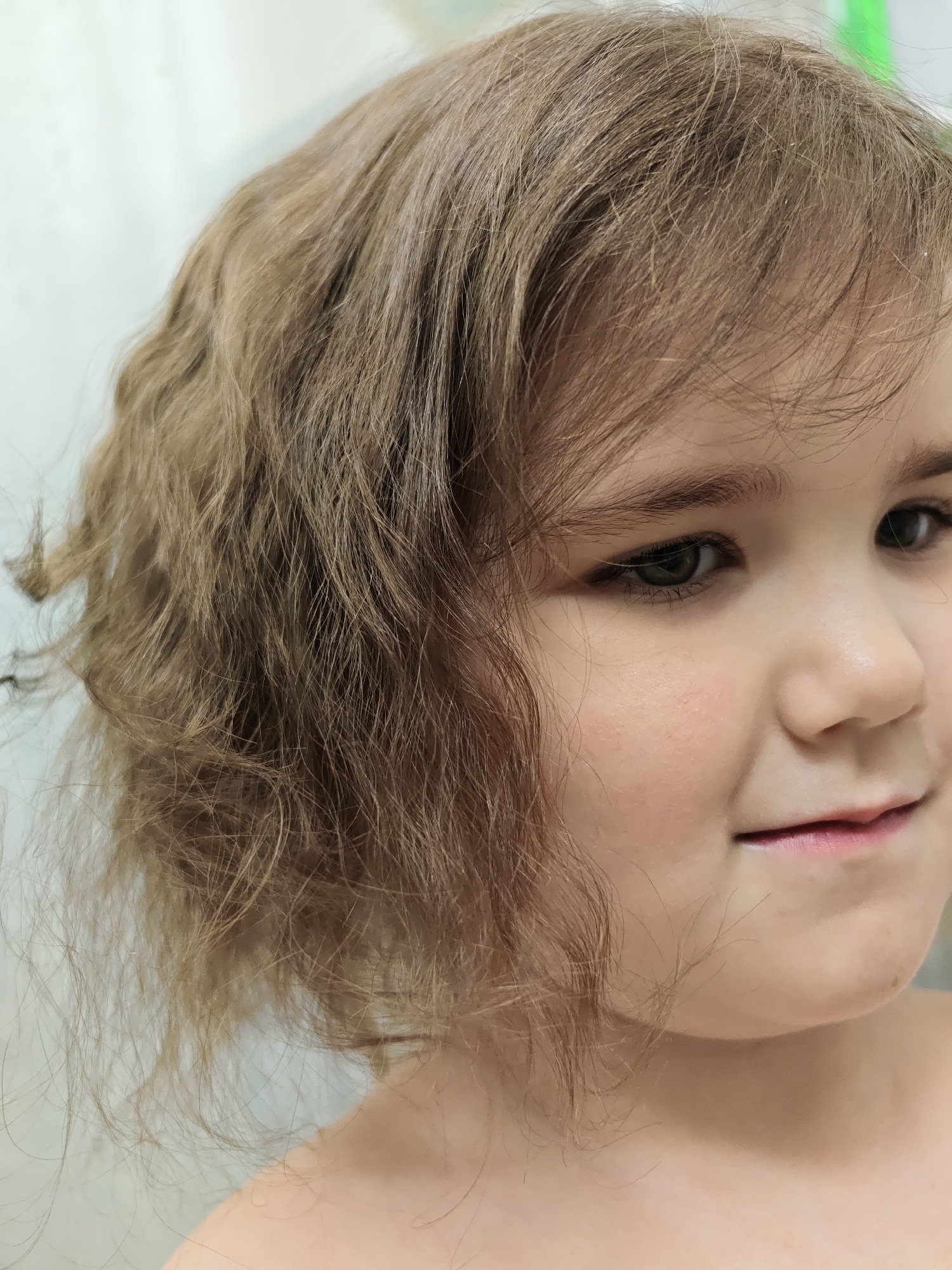 A close-up, vertical portrait captures the right side of a young girl's face. She has light brown, shoulder-length hair with noticeable waves and curls, which appears slightly disheveled with individual strands visible, some showing a finer texture around the temples. Her fair complexion shows a subtle rosy blush on her cheeks. She possesses dark, expressive eyes that gaze slightly upward and to the viewer's left, framed by dark eyebrows and eyelashes. A gentle, closed-lip smile is present on the visible corner of her mouth. Her bare shoulders and upper chest are visible, suggesting she is undressed or wearing minimal clothing, possibly indicative of a bath time setting or a casual indoor moment.
The background is softly blurred and light-colored, hinting at an indoor, domestic setting. A faint, indistinct green blur can be seen in the upper right corner of the background. The child's expression conveys a sense of calm and mild curiosity or perhaps a hint of shyness. The photograph appears to be taken indoors, likely during the daytime or with bright artificial light. No specific activity beyond being photographed is depicted. No visible text is present. The geographical context for this scene is Chaikovsky, Russia.