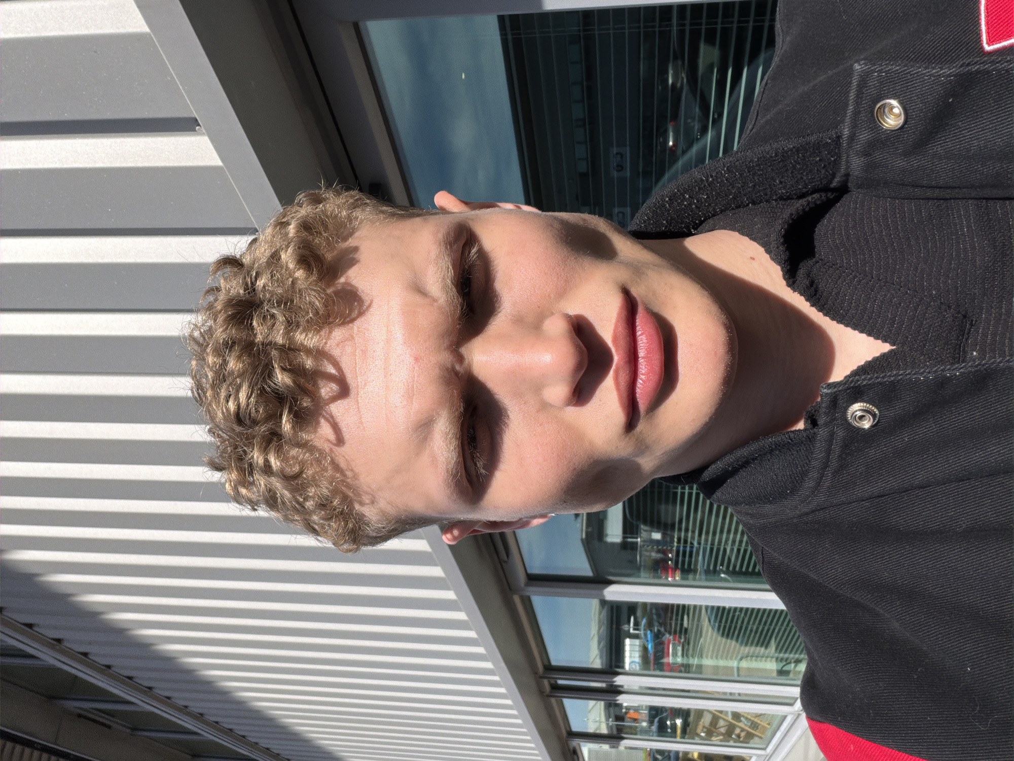 A close-up, rotated image depicts a fair-skinned young man with tightly curled blond hair, clad in a black jacket, against a backdrop of what appears to be a commercial building with glass windows and white horizontal metal or plastic paneling. Reflections in the windows suggest an exterior environment possibly containing vehicles. The lighting is bright and sunny, casting strong shadows that emphasize the texture of the building materials and the subject's facial features. There is insufficient detail in the image to specifically verify the location in Trafford, United Kingdom.