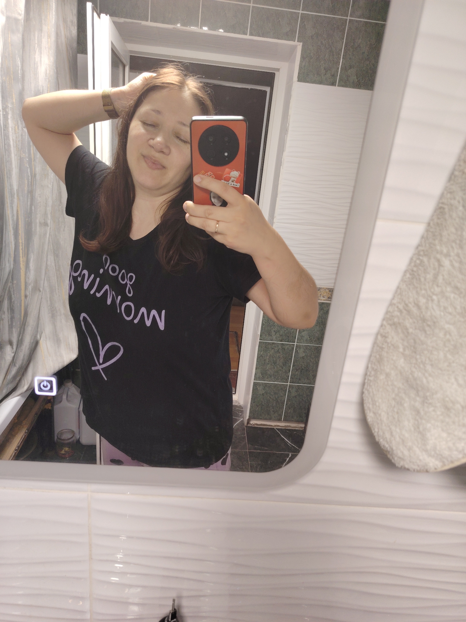 A woman is taking a selfie in a bathroom. She is wearing a black t-shirt with purple lettering that reads "good morning" and a heart. She has her arm raised, touching her hair. The background shows a mirror, a tiled wall, and a shower curtain. The mirror reflects the woman, a phone, and items on a shelf. A towel hangs to the right of the mirror. The scene suggests a personal moment, likely in a residential bathroom.