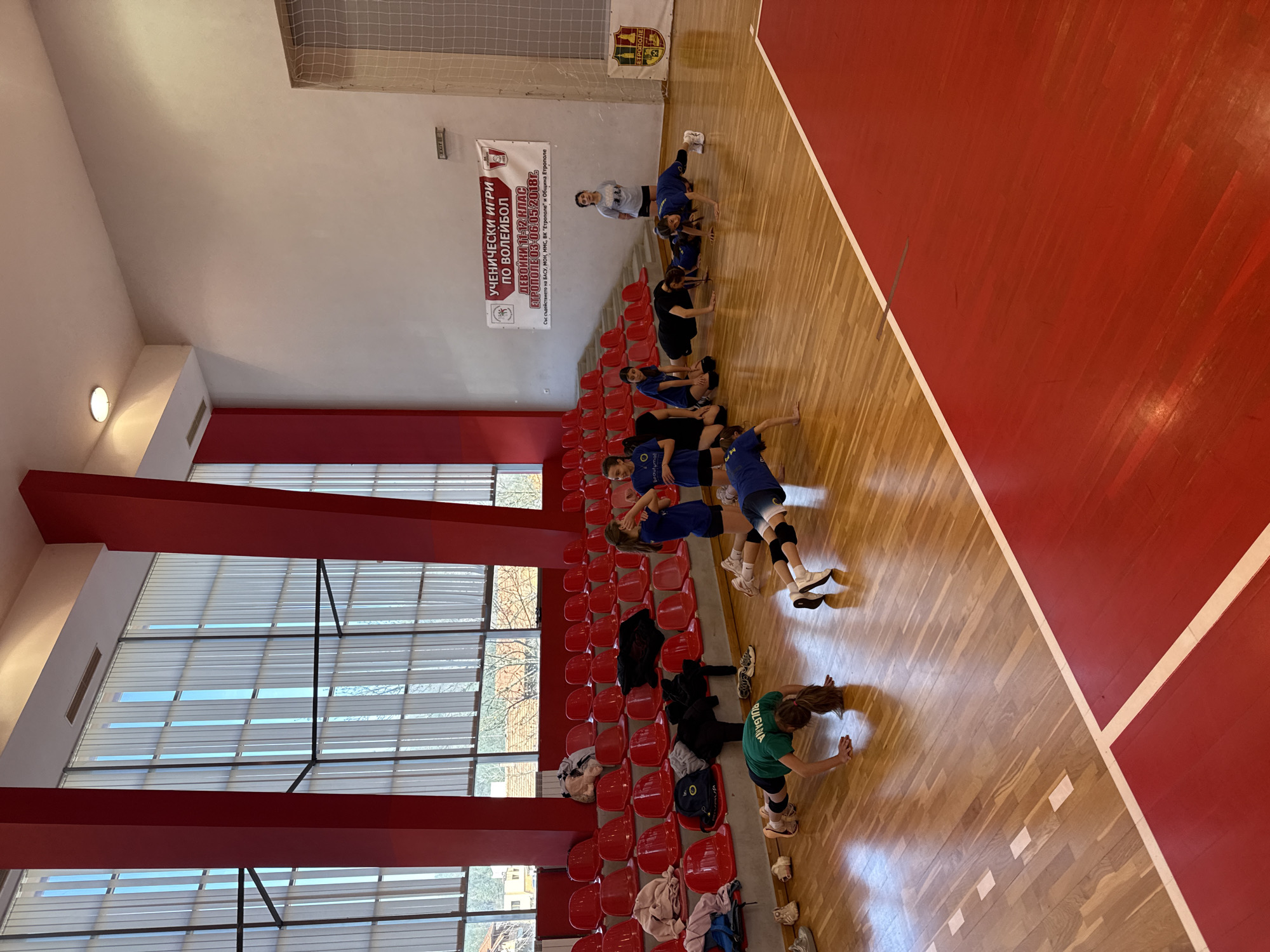 The image shows a group of young women, likely a volleyball team, stretching or warming up in a gymnasium or sports hall. They are near rows of red seats and a large window. In the background, there is a banner with the text "УЧЕНИЧЕСКИ ИГРИ ПО ВОЛЕЙБОЛ" (School Games in Volleyball) and "ЕТРОПОЛЕ 03-06.05.2018г." suggesting the location is Etropole, Bulgaria, during a volleyball event held between May 3rd and 6th, 2018. The team members are wearing athletic apparel, and their actions suggest they are preparing for a game or practice. The gymnasium has a wooden floor with red painted sections indicating the boundaries of the court.