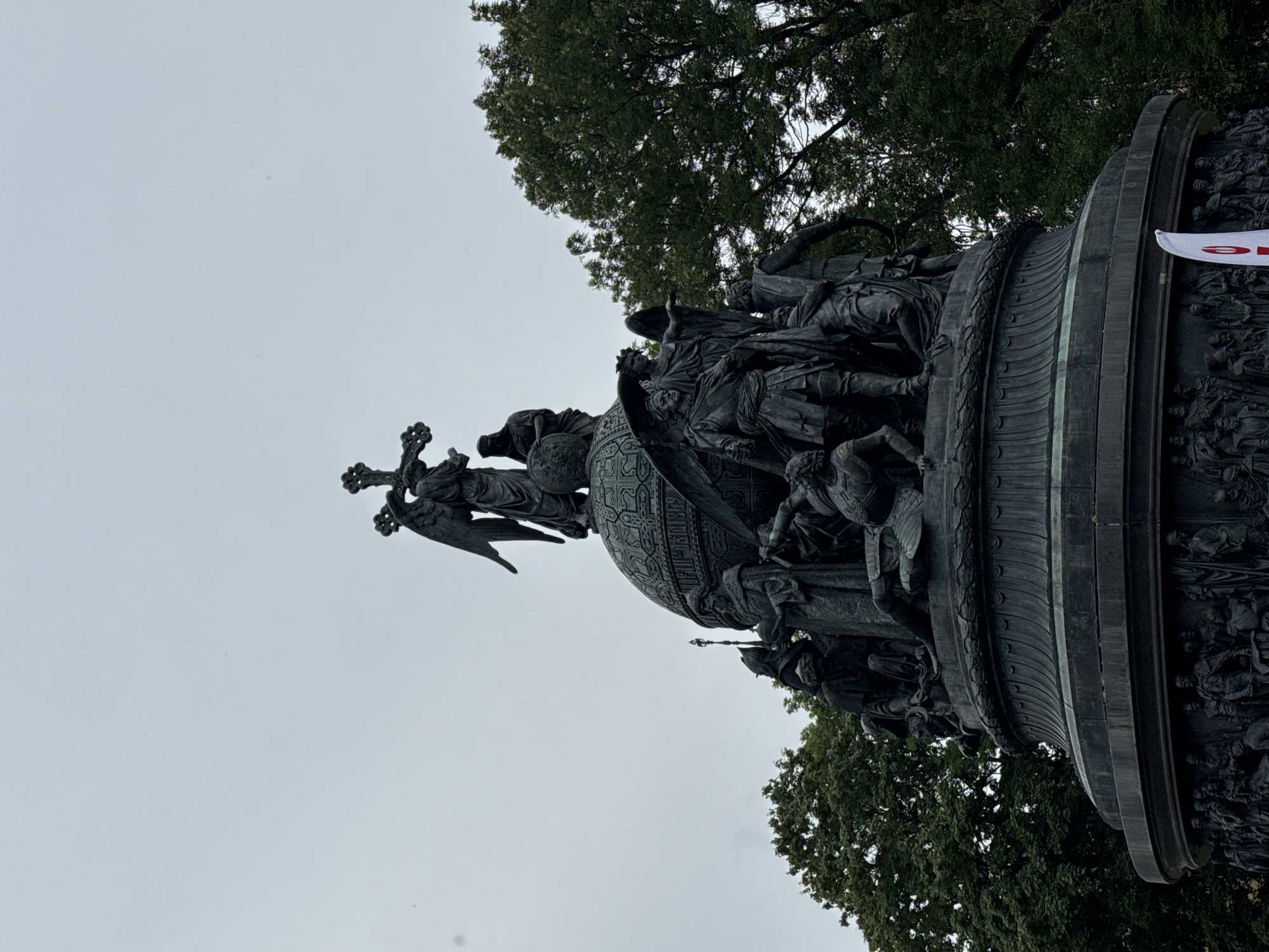 This is a low-angle, full shot of the Millennium of Russia monument, located in Veliky Novgorod, Russia. The monument is made of dark bronze and depicts numerous figures in relief, as well as larger sculptural elements. At the very top, a winged angel figure holds a cross. Below the angel, an ornate globe is surrounded by figures representing different historical eras and important individuals in Russian history. The monument is set against a pale, overcast sky with the tops of green trees visible on the right and left sides of the frame. The base of the monument has a detailed frieze of smaller figures. A white flag with a red symbol is partially visible in the lower right corner. The overall impression is one of historical significance and artistic grandeur.