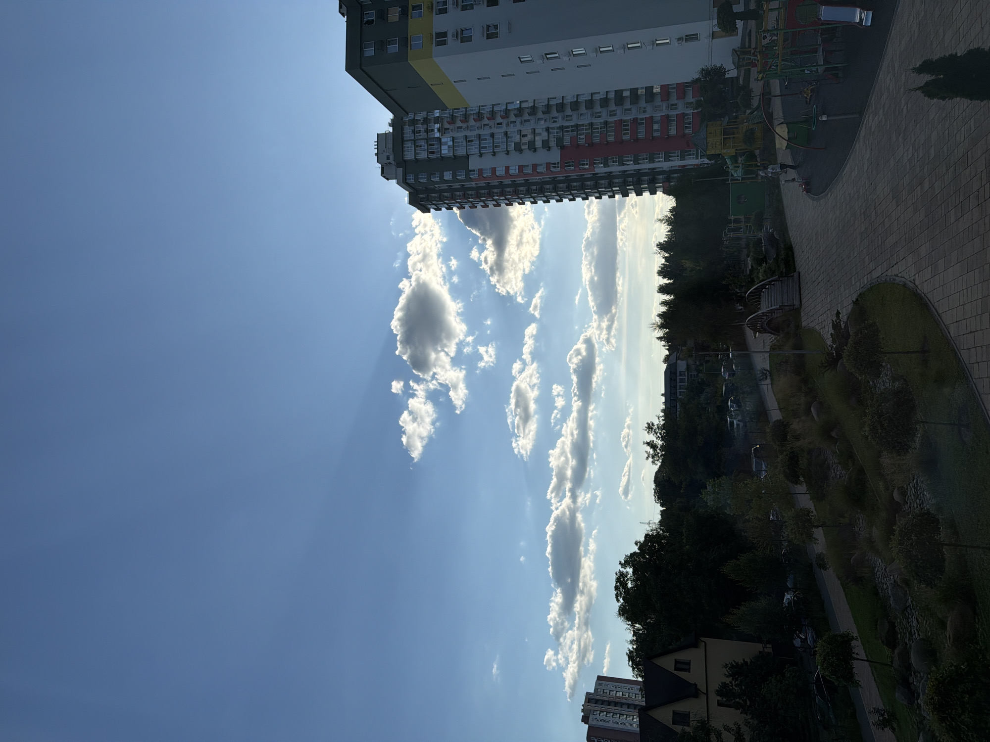 The image captures a residential area with modern high-rise buildings, a playground, and a park-like landscaping in Kyiv, Ukraine. The sky is blue with scattered white clouds, and sun rays are visible piercing through the clouds. In the foreground, a paved pathway winds through a landscaped area with rocks, small trees, and bushes. To the right, a colorful playground with slides and swings is visible. In the background, a tall residential building with a gray and red facade stands prominently, with another building to its right that is mostly white and yellow. Further in the distance, there are more buildings and trees. The scene appears to be set during the day, possibly late afternoon, given the lighting. There are a few people visible in the playground area.