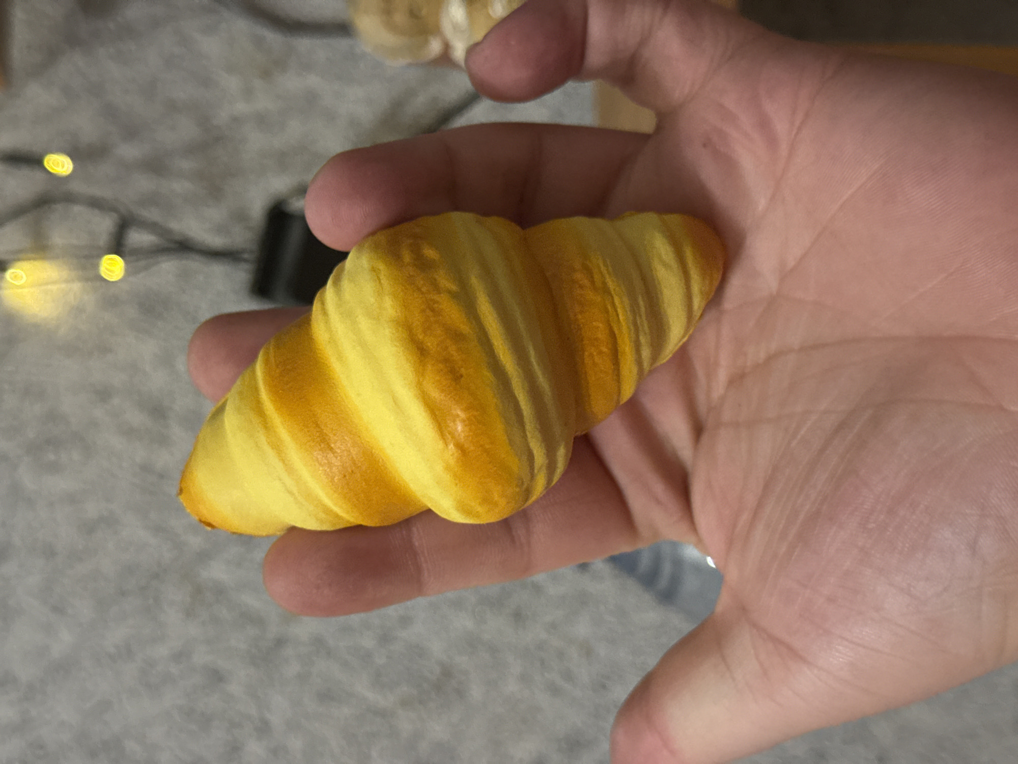 An over-the-shoulder, close-up shot shows a person's left hand holding a miniature croissant-shaped toy. The toy is a bright yellow with orange swirls mimicking the flaky layers of a real croissant. The background is out of focus, but appears to be an indoor setting with a textured gray surface, possibly carpet, and out-of-focus string lights. There are no visible landmarks or indicators of a specific location within Molodezhnyi, Russia.