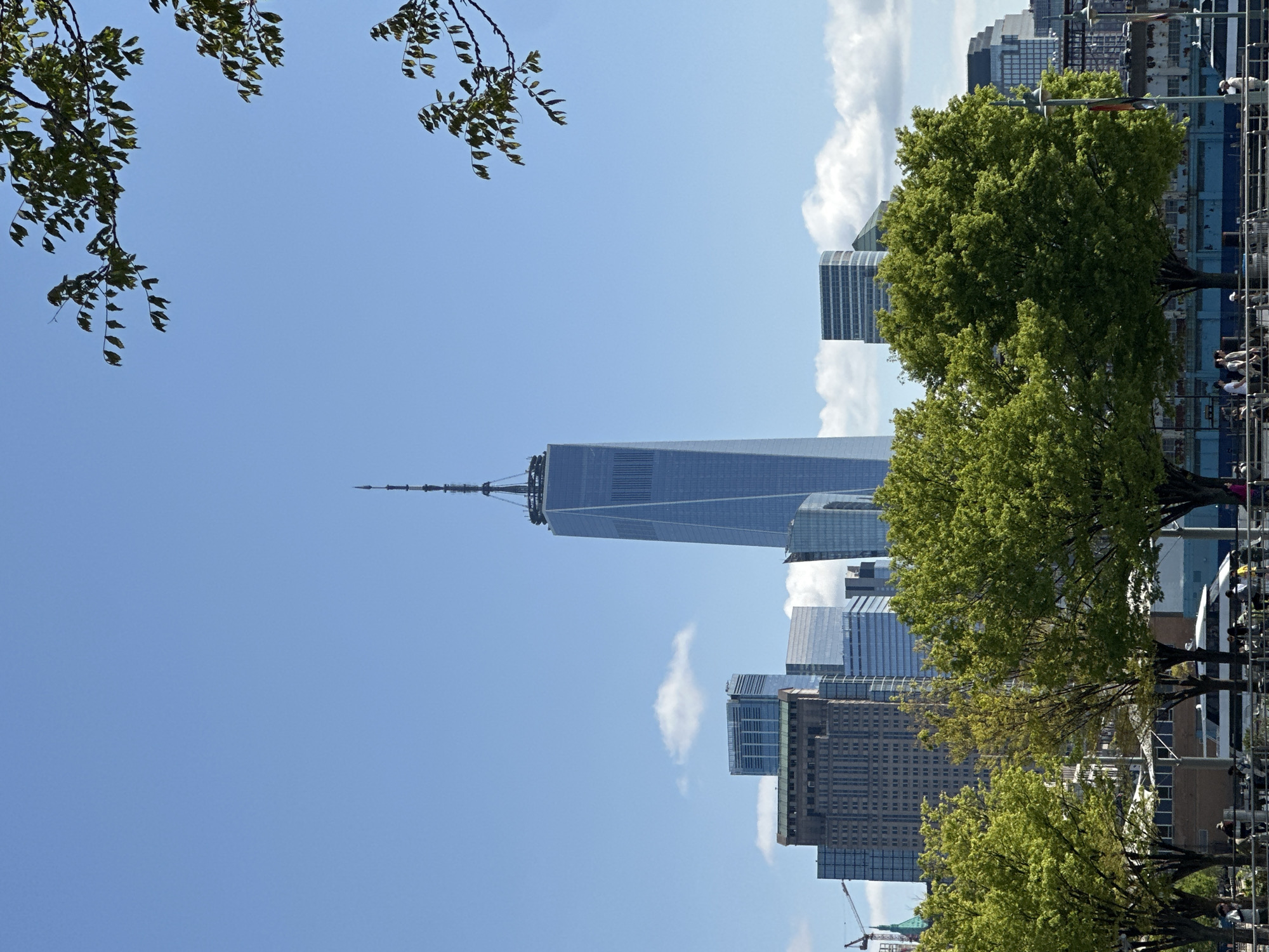 The image shows a view of the New York City skyline in the United States. The focus is on the One World Trade Center building, which stands prominently. The foreground includes green trees and foliage. Some clouds are in the blue sky. Other buildings are visible, and a section of a crane is present in the bottom left corner. In the distance, people can be seen.