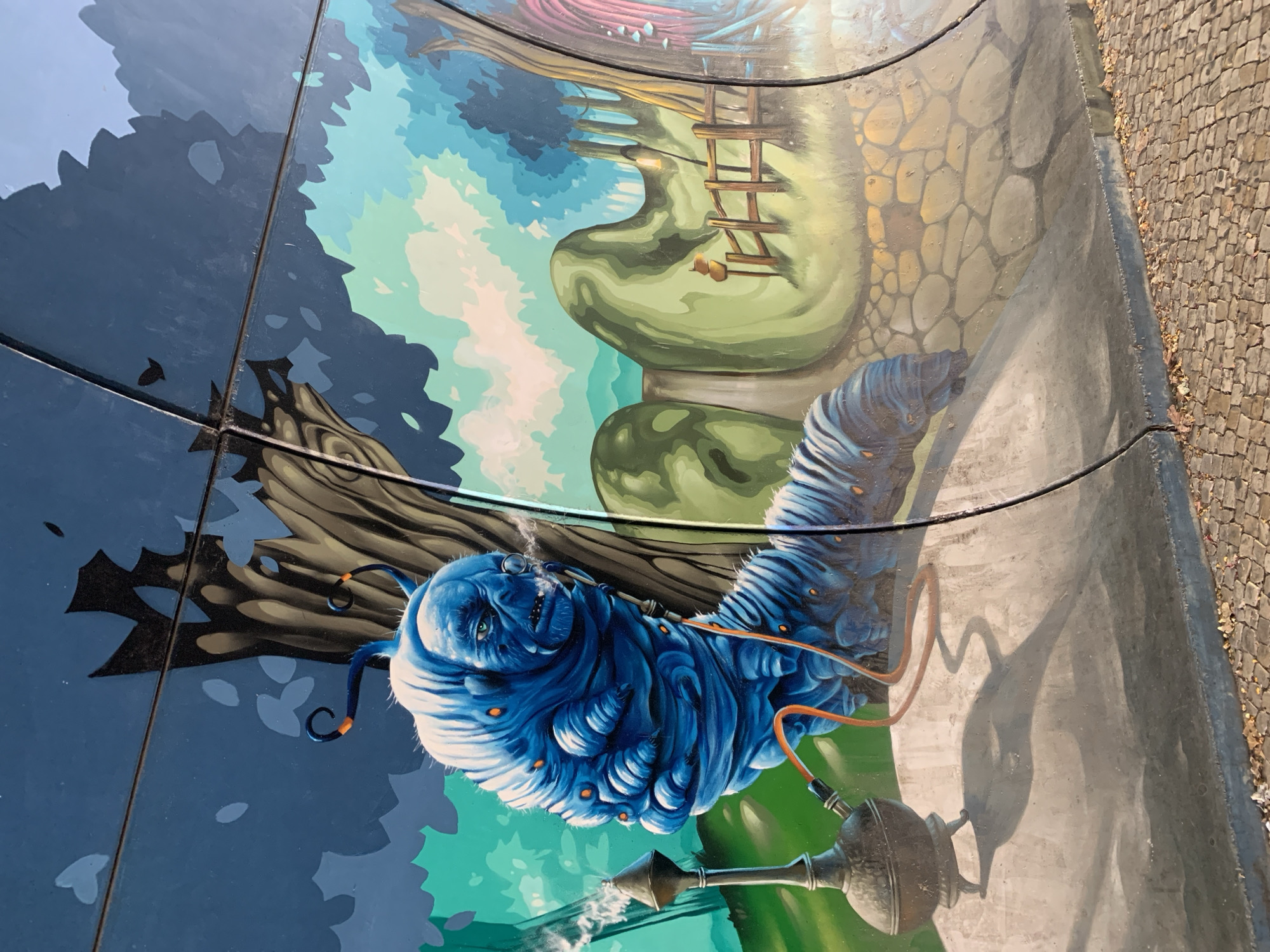 This media file depicts a mural painted on a curved surface, likely a wall or a structure. The artwork features a large, blue, anthropomorphic caterpillar with distinct facial features, including wide eyes and a somewhat smug expression. The caterpillar is shown smoking from a hooka pipe, with smoke emanating from it. An orange tube connects the hooka to a nozzle near the caterpillar's mouth. The caterpillar is depicted against a surreal landscape with large, rounded green shapes that could represent oversized foliage or strange rock formations. A wooden ladder leans against one of these green shapes. The background transitions from a dark blue and black area, suggestive of a night sky or shadowy trees, to lighter blue and green hues, hinting at a dreamlike or fantastical environment. The artwork is situated outdoors, as evidenced by the stone-paved ground visible on the right side, some fallen leaves, and the natural lighting. The time of day appears to be daytime, likely sunny, given the shadows cast. The style of the mural is illustrative and detailed, with a vibrant color palette. The location context of Odivelas, Portugal, suggests this is a public art installation. There is no visible text within the media file.