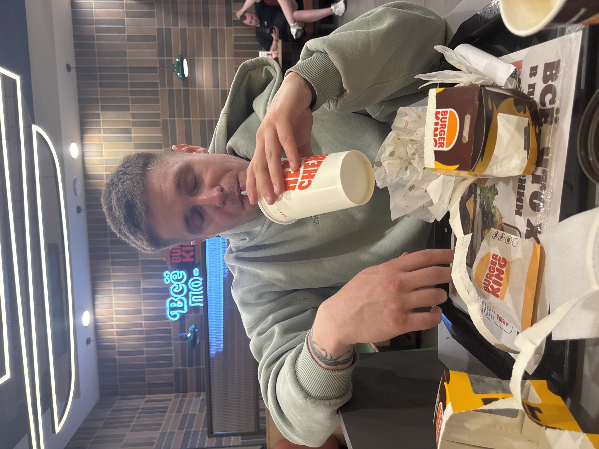 A man in a light green hoodie is sitting at a Burger King restaurant table, drinking from a large paper cup with a straw. On the tray in front of him are two partially eaten burgers and a box of fries. The restaurant appears to be located in Sochi, Russia, based on the Cyrillic script on the tray liner and a neon sign reading "Всё по-сочински" (Everything in Sochi style). Other patrons can be seen in the background.