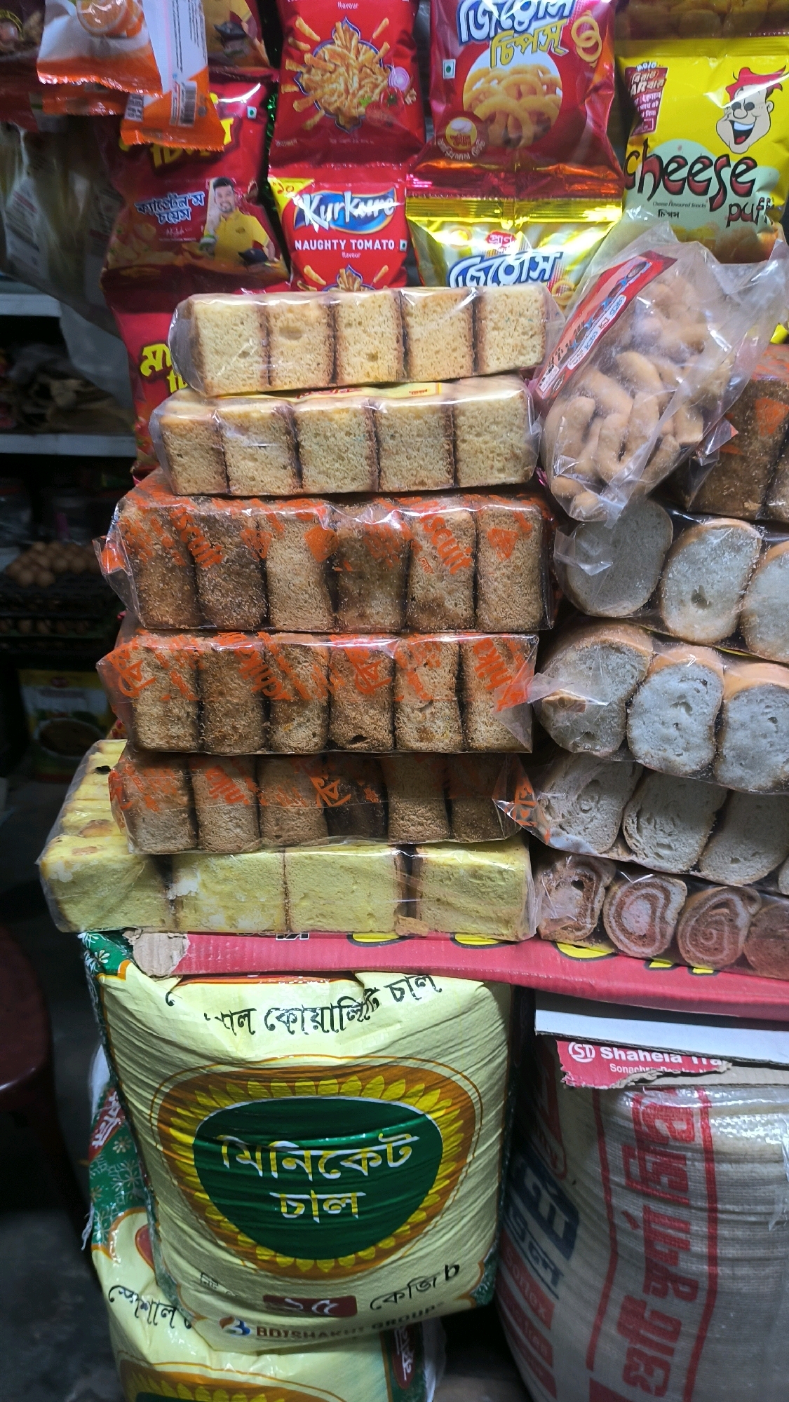 A small-scale retail shop in Begumganj Upazila, Bangladesh, displays a variety of packaged food items, primarily snacks and baked goods. The foreground features stacks of plastic-wrapped slices of what appears to be rusk or sponge cake, some with a slightly toasted appearance. To the right, a bag of assorted fried snacks is visible, along with packaged bread slices. Behind these, several colorful snack packets, including brands like "Kurkure Naughty Tomato" and "Cheese Puff," are arranged. In the lower portion of the image, a large, yellow bag with green and yellow branding, possibly containing lentils ("Dal"), is prominent. The overall environment suggests a typical local market setting with goods stacked for sale. No people are visible in the frame.