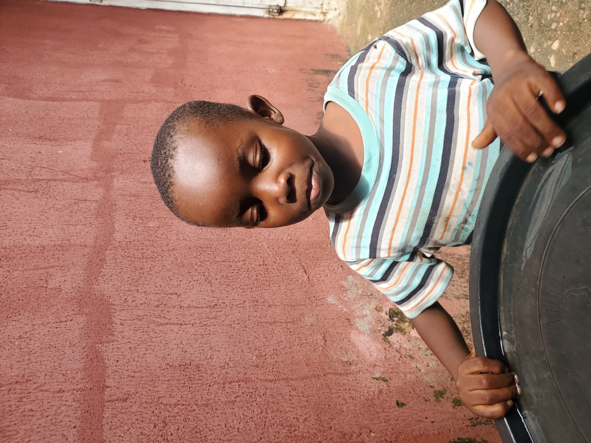 In Jos, Nigeria, a young Black boy with a shaved head and downcast eyes stands against a textured, reddish-pink wall. He wears a horizontally striped t-shirt with bands of white, light blue, navy, and orange. His hands are on the rim of a large, black plastic container, which appears to be filled with water. The container is positioned in the foreground, partially obscuring the boy's lower body. The wall behind him shows signs of wear and tear, with patches of peeling plaster and greenish discoloration in the lower left corner. To the right of the wall, a white door or shutter is visible. The ground beneath the boy is paved with concrete. The lighting suggests it is daytime, with sunlight casting subtle shadows.