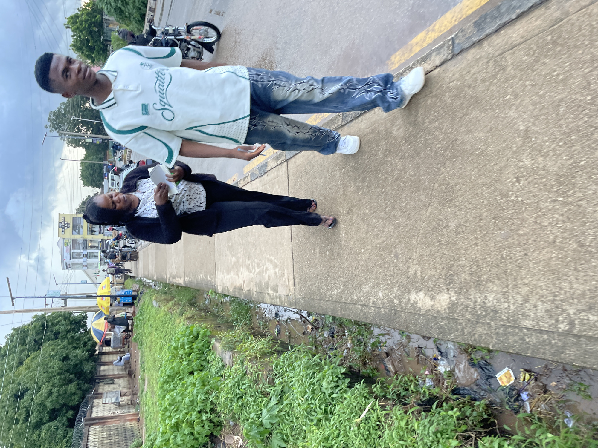The image depicts a street scene in Jos, Nigeria, likely during daylight hours, with overcast or cloudy conditions. Two individuals are walking on a concrete sidewalk. The person in the foreground on the right is a young male wearing a white polo shirt with green trim and denim jeans adorned with a flame-like pattern. He is looking slightly towards the camera. Beside him, to the left and slightly behind, is a woman wearing a dark blazer over a patterned blouse and dark trousers. She is looking down at a paper or book in her hands.
The sidewalk runs parallel to a street where several motorcycles and vehicles are present, suggesting a moderately busy urban environment. On the left side of the sidewalk, there is a drainage ditch filled with murky water and debris, overgrown with lush green vegetation. Further down the street, market stalls with umbrellas are visible, and other pedestrians are present. Buildings line the opposite side of the street, some appearing to be shops or commercial establishments. The overall impression is of an everyday urban scene in a developing city.