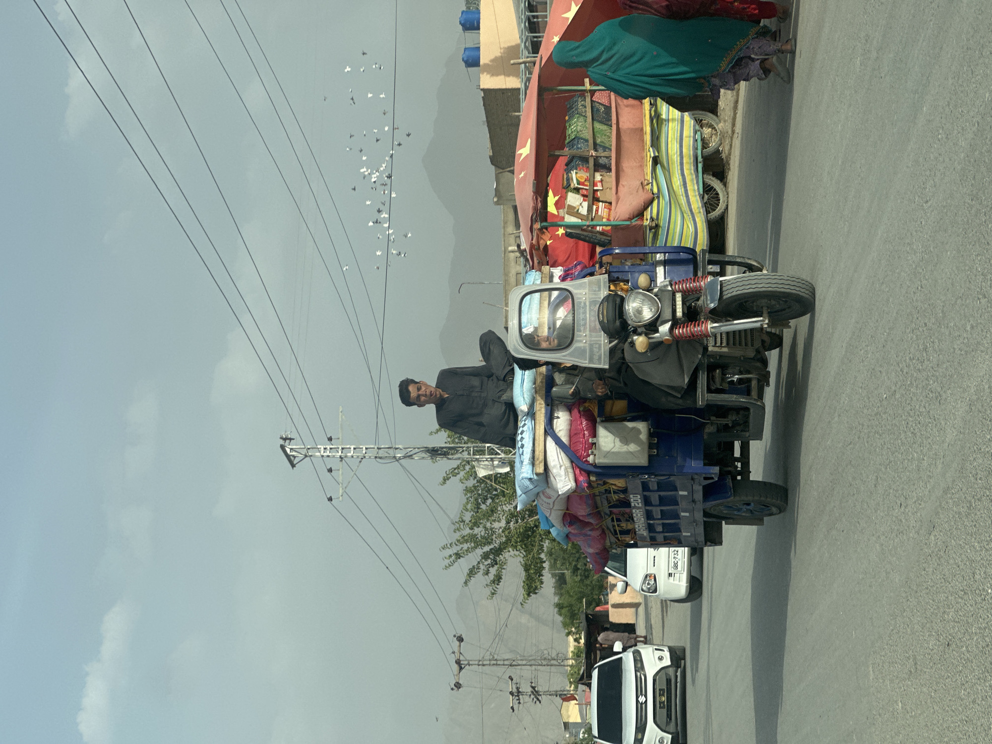 The image captures a street scene in Quetta City Tehsil, Pakistan, featuring a man driving a blue three-wheeled vehicle loaded with goods. The vehicle is positioned on the right side of the road, heading forward. The man is wearing a dark-colored shirt and is facing the viewer. The vehicle is full of various items, including sacks, pillows, and other belongings. In the background, other vehicles are present, including a white sedan and a three-wheeled vehicle with a red awning, which seems to be a small shop. Overhead, power lines traverse the sky. A flock of birds is visible in the upper left corner. Mountains can be seen in the distance. The weather appears overcast with a grey sky.