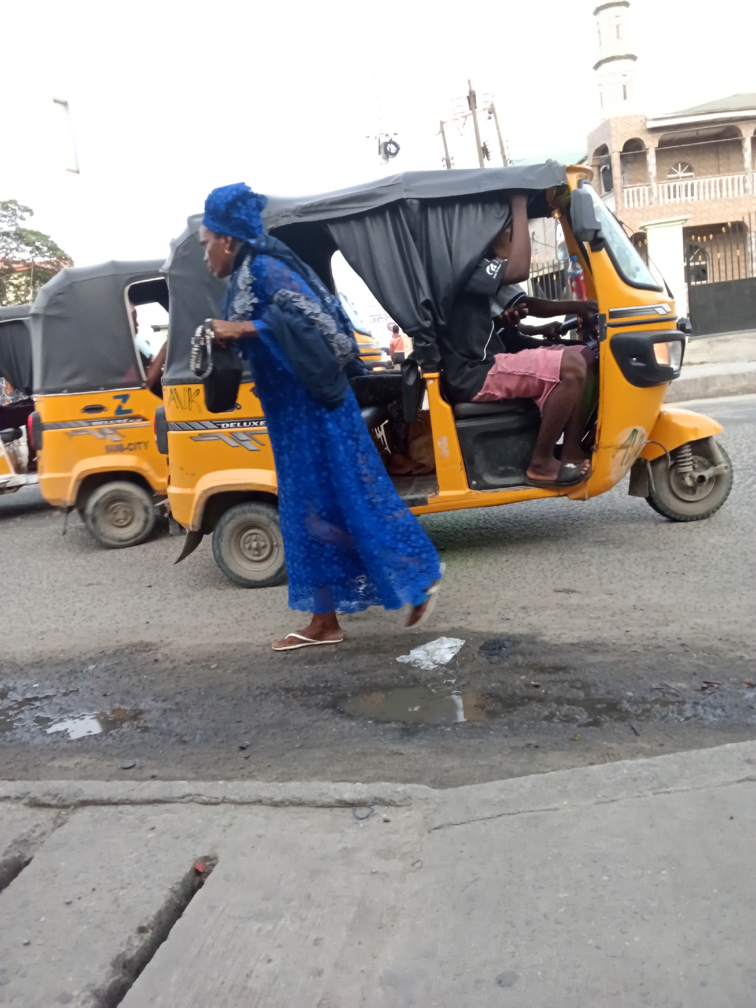 A daytime street scene in Ado, Nigeria, features a woman walking alongside two yellow auto-rickshaws (keke napeps). The woman, dressed in a long blue, lace-patterned abaya or dress and a matching head covering, is seen in profile, moving from left to right. She carries a black bag with a silver-colored chain strap in her right hand and wears light-colored flip-flops. Her left foot is lifted mid-stride, suggesting movement.
To her right, a yellow auto-rickshaw with a black canopy is prominently featured. Inside, a person, likely the driver, is seated, wearing a black top and pink shorts, with their hands on the handlebars. A red bottle is visible inside the vehicle. The word "DELUXE" is clearly visible on the side of this auto-rickshaw, along with partial letters "ANK" near the front wheel.
Behind the first auto-rickshaw, a second one, also yellow with a black canopy, is partially visible. Text on its side includes "SUB-CITY" and what appears to be a "Z". The auto-rickshaws are positioned on a paved road that shows signs of wear, including potholes and puddles, suggesting either recent rain or poor drainage. A concrete gutter with a red object (possibly a cap) lies in the foreground.
In the background, under an overcast sky, a multi-storied building with distinctive arched windows, balconies, and a minaret-like structure is visible, hinting at a mosque or Islamic architectural influence in the area. The overall scene captures a typical urban moment with local transportation and pedestrian activity.