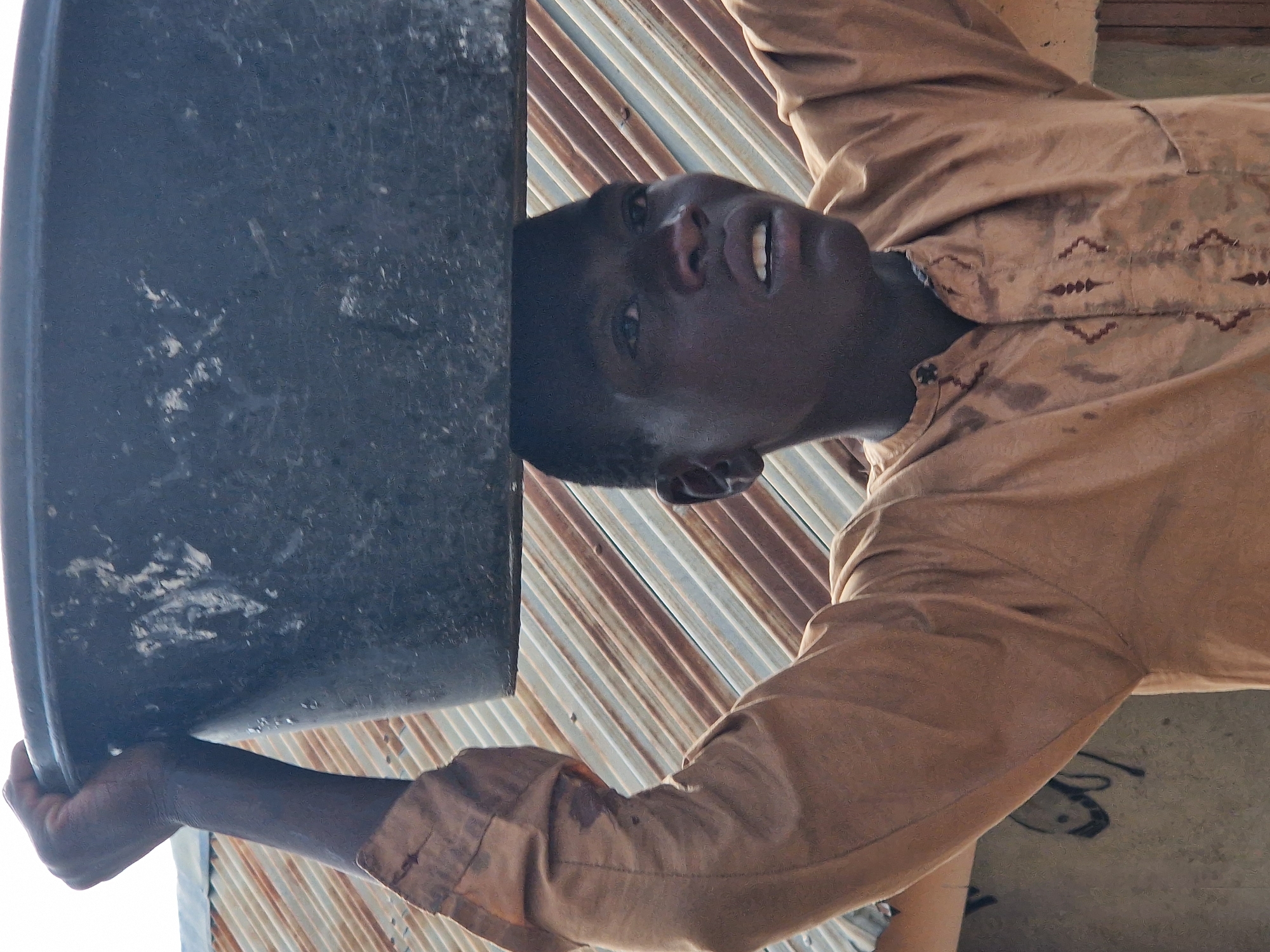 A young Black person, appearing to be a male, is shown from the chest up in a medium shot. The individual is wearing a tan, collared garment with decorative stitching down the front. They are carrying a large, dark-colored plastic basin on their head with their right arm raised to support it. The background consists of a corrugated metal roof with alternating white and rusty brown panels, suggesting an outdoor environment. The person's facial expression is one of exertion or concentration. The lighting suggests it is daytime, with a slight shadow cast on the person's face from the basin.