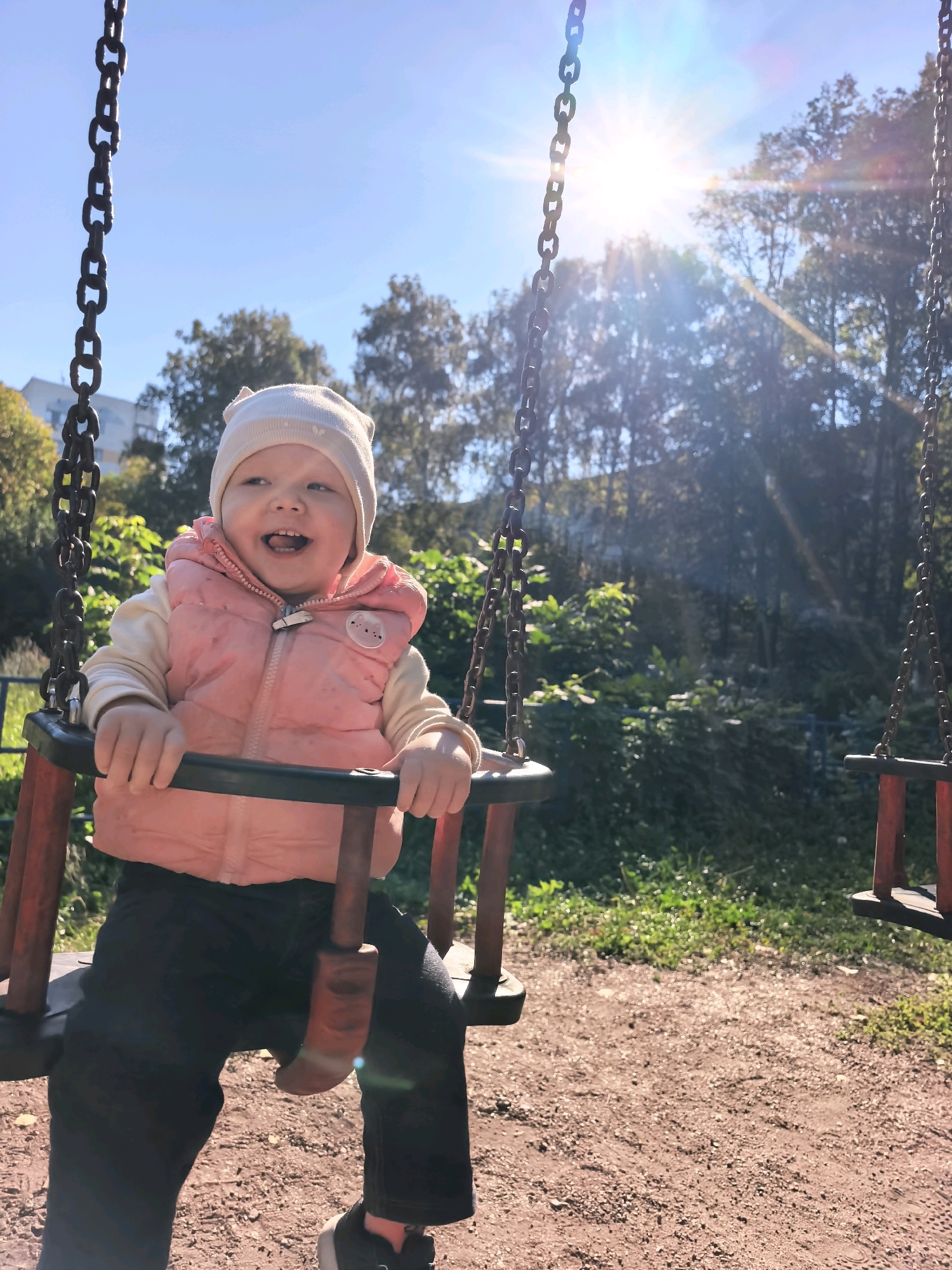 A young child, likely a toddler, is seated on a swing at a playground. The child is wearing a light-colored hat and a pink vest over a cream-colored long-sleeved shirt, with dark pants and black sneakers. The child has a joyful expression, with their mouth open and eyes looking towards the right. The swing set has sturdy metal chains and wooden seats. The playground appears to be located outdoors, with a background of trees and green foliage. The sun is shining brightly, casting lens flares and dappling the scene with light. The ground beneath the swing is covered in brown dirt and small stones. The overall setting suggests a sunny day at a public park or playground in Sterlitamak, Russia.