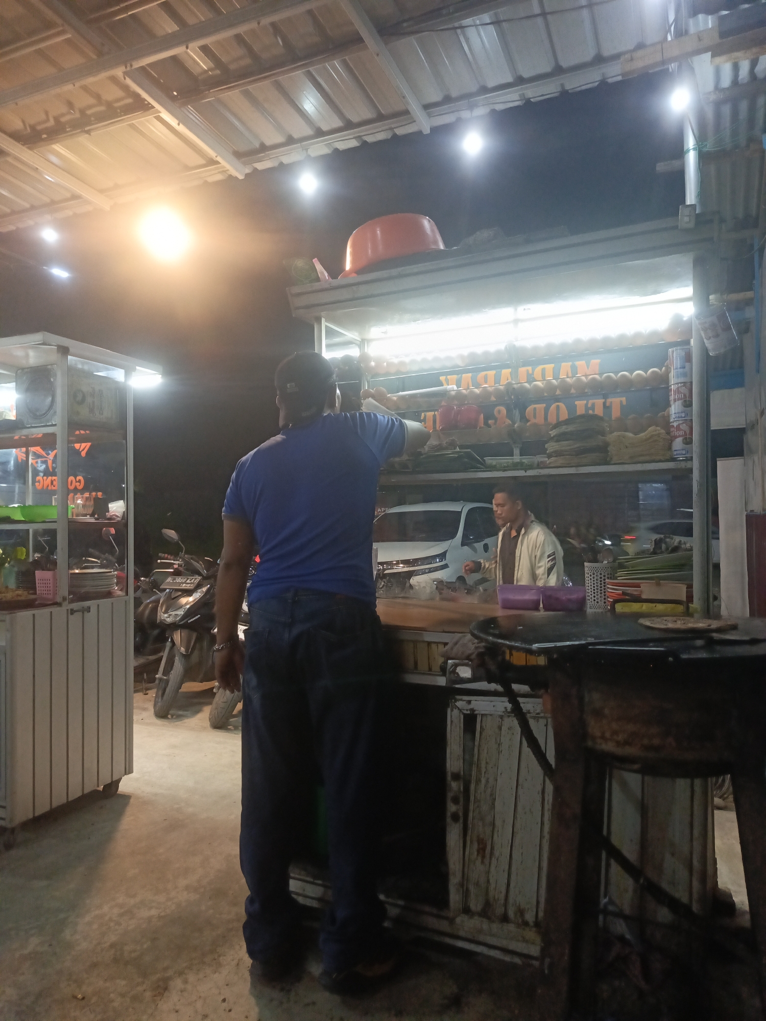 A street food stall is operating at night, possibly in Lhokseumawe, Indonesia. The stall is illuminated by bright overhead lights. A man in a blue t-shirt and jeans stands with his back to the camera, facing the stall. Another man in a light-colored jacket is visible behind the counter, interacting with customers. The stall is stocked with food items, including what appear to be fried snacks, and has a display of plates and other serving items. A motorcycle is parked near the stall. The ground is concrete, and the overall scene suggests a busy, informal commercial environment.