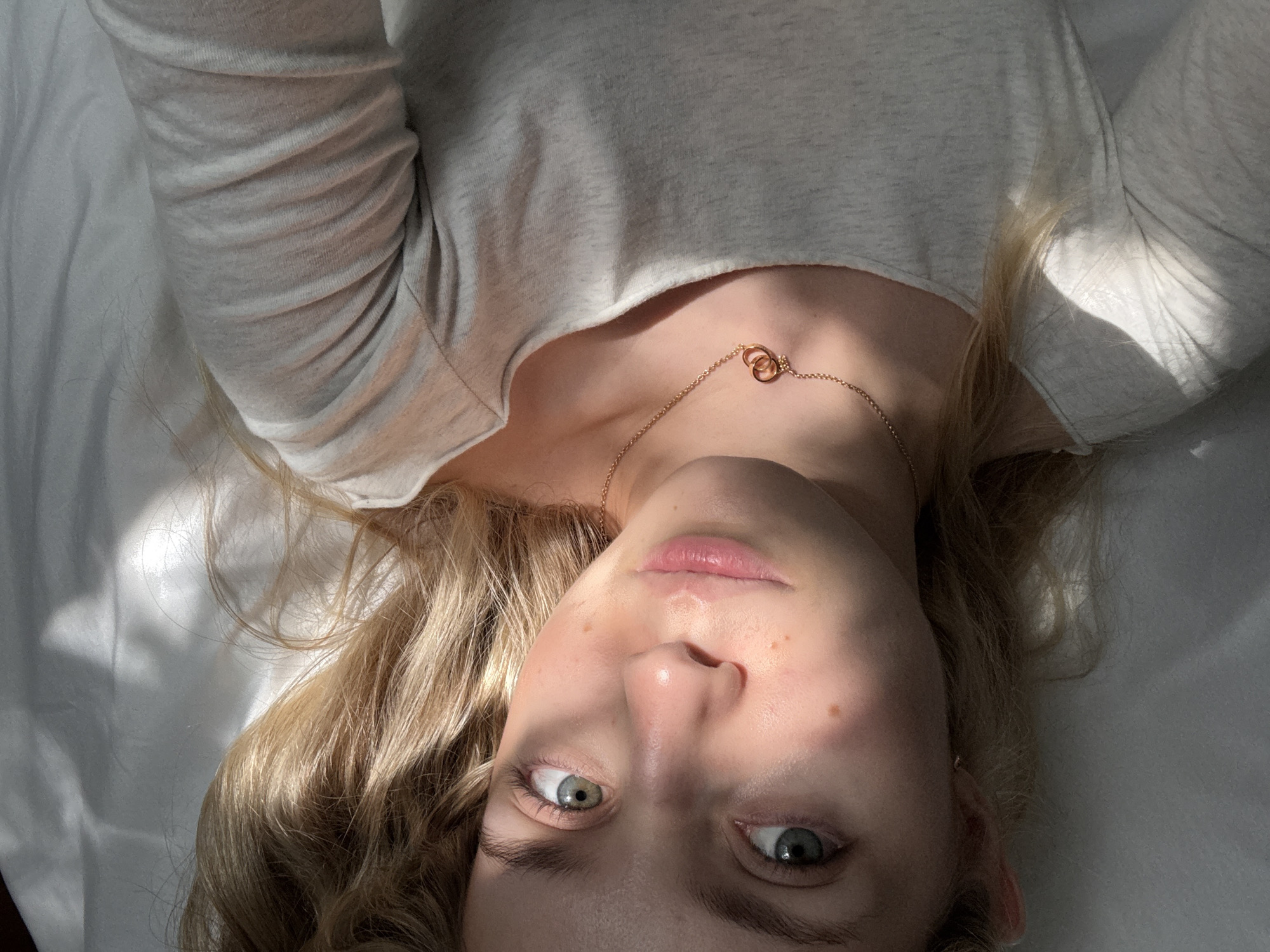 A young woman with blonde hair and light green eyes lies on her back on a white surface, likely a bed or sofa. She is wearing a light gray long-sleeved shirt and a delicate gold necklace with two interlocked rings. Sunlight streams across her face, highlighting her features and casting shadows on the fabric of her shirt. Her hair is spread out around her head. The image appears to be a close-up, self-taken photograph. There is no discernible indication of a specific location within the city of Miami, United States of America.
