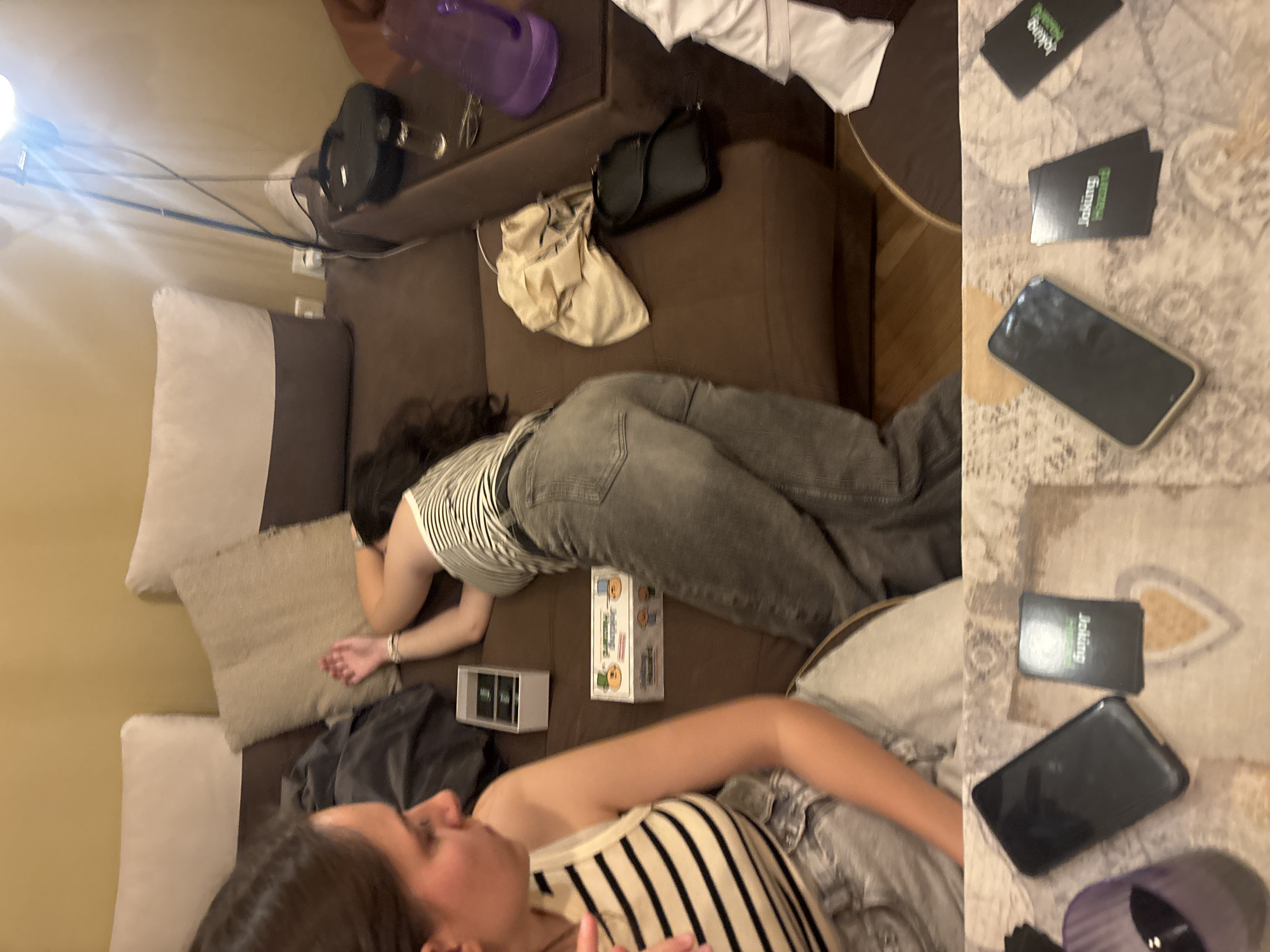 The image captures an indoor scene, likely a living room, where two young women are relaxing on a brown sofa. The picture is rotated counter-clockwise, showing the scene from an overhead perspective.
One woman is lying face down on the couch, wearing a striped top and grey jeans, her head turned away from the camera. Her arms are bent, suggesting a relaxed posture. Near her, on the couch, is a white box with cartoon-like characters and the partially visible text "Joking Hazar...", indicating it's likely the card game "Joking Hazard." A grey/silver rectangular object is also visible nearby.
In the foreground, partially visible, another woman lies on her back, also wearing a striped top. Her face is mostly out of frame.
Various objects are scattered around the scene, suggesting a casual gathering. On the armrest or a nearby surface, there's a black handbag, a light-colored fabric bag, and a purple water bottle or cup. A black speaker or electronic device is also present on an elevated surface.
To the right, on what appears to be a patterned rug or blanket on a table, several black cards with prominent green text reading "Joking" are laid out. Two smartphones, one black and one silver, are also on the rug, along with another purple item, possibly a cap from the water bottle.
The background shows a light-colored wall with visible power sockets and a floor lamp emitting a bright artificial light, indicating it might be evening or night. The overall atmosphere is one of comfort and relaxation, possibly after or during a casual game night. There are no specific cues within the image itself to suggest the location is Plovdiv, Bulgaria, but the setting is a typical modern home interior.