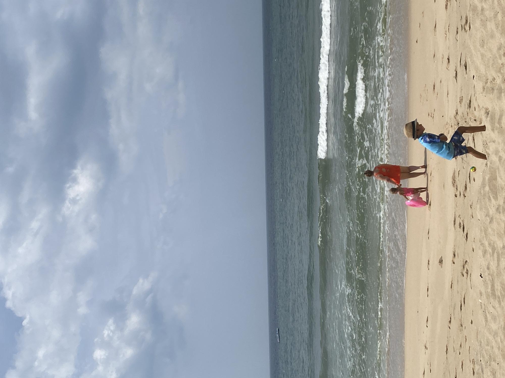 The image depicts a sandy beach with the ocean and a cloudy sky. In the foreground, a young boy wearing a blue shirt and patterned shorts, with a straw hat, is playing with a string toy. He is facing away from the camera and to the left, with his body crouched slightly. To the left of the boy, a woman in an orange top and a young girl in a pink swimsuit holding a pink inflatable ring are standing near the water's edge. The woman is facing away from the camera, and the girl is facing towards the ocean. Gentle waves are breaking on the shore. In the distance, on the horizon, a small boat is visible. The beach is composed of light-colored sand with scattered footprints and small mounds. The ocean appears green and blue under a gray, overcast sky. The scene is consistent with a public beach in Mũi Né, Vietnam, a coastal city known for its beaches and water activities.