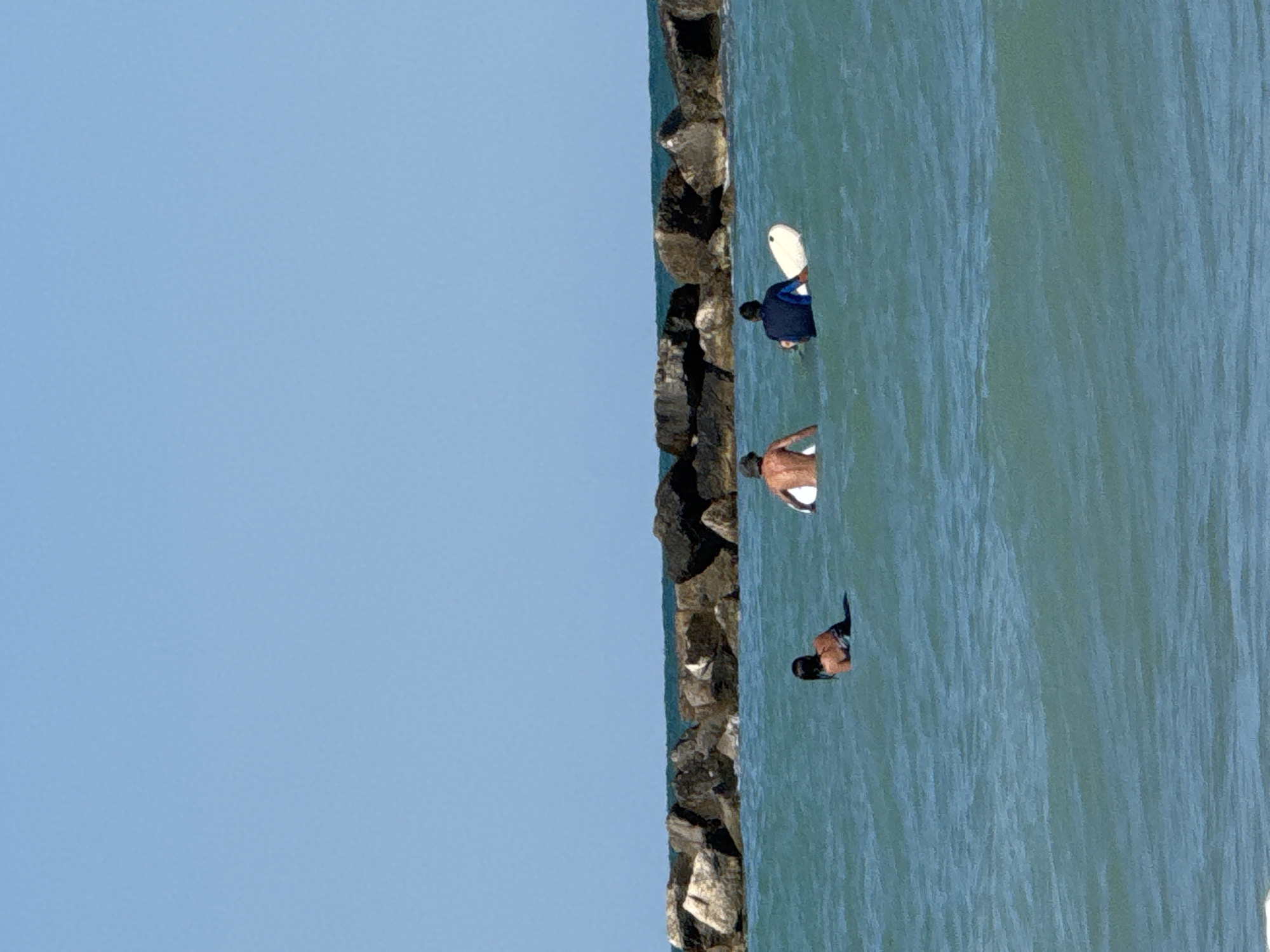 Three people are in the turquoise waters of the Adriatic Sea, near San Benedetto del Tronto, Italy. A rock breakwater runs across the middle of the image, separating the calmer waters in the foreground from the choppier sea beyond. The sky is clear and a bright blue.
On the left, a person with dark hair is partially submerged in the water, looking towards the left. In the center, a person with light skin, possibly male, is sitting on a surfboard, facing away from the camera, with their body partially visible. To the right, another person, also facing away, is sitting on a white surfboard with a blue shirt. They appear to be preparing to surf or have just finished. The water is clear and shows some ripples.