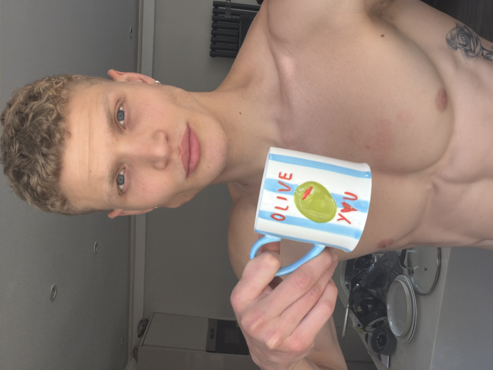 A fair-skinned man with blonde curly hair and blue eyes is holding a mug that says "Olive You." He is shirtless and has a tattoo of a rose on his right side. The man is in what appears to be a kitchen, with cabinets and a sink visible in the background. The lighting suggests it is daytime. The man's expression is neutral. The image is cropped to focus on the man and the mug. The specific location in London, United Kingdom cannot be determined from the image.