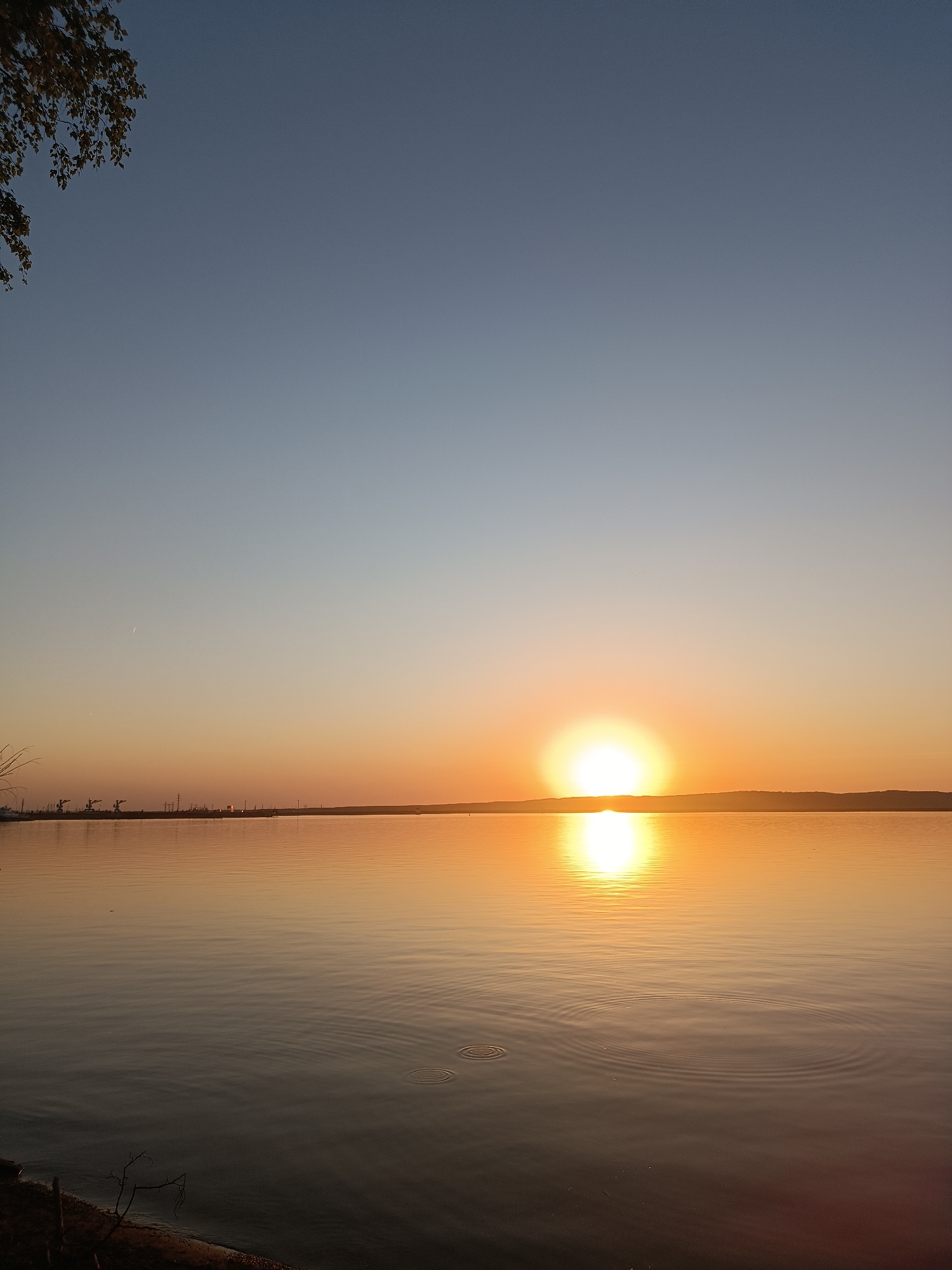 The image shows a sunset over a body of water, possibly a lake or wide river, in Chaikovsky, Russia. The sun is positioned just above the horizon, creating a bright reflection on the water's surface. The sky transitions from a light blue at the top to a warmer orange and yellow near the horizon. There is an industrial area visible on the far left, with cranes. There are trees in the upper left corner and in the lower left corner. There are ripples on the water surface. The lower part of the image is dark, depicting the shoreline.