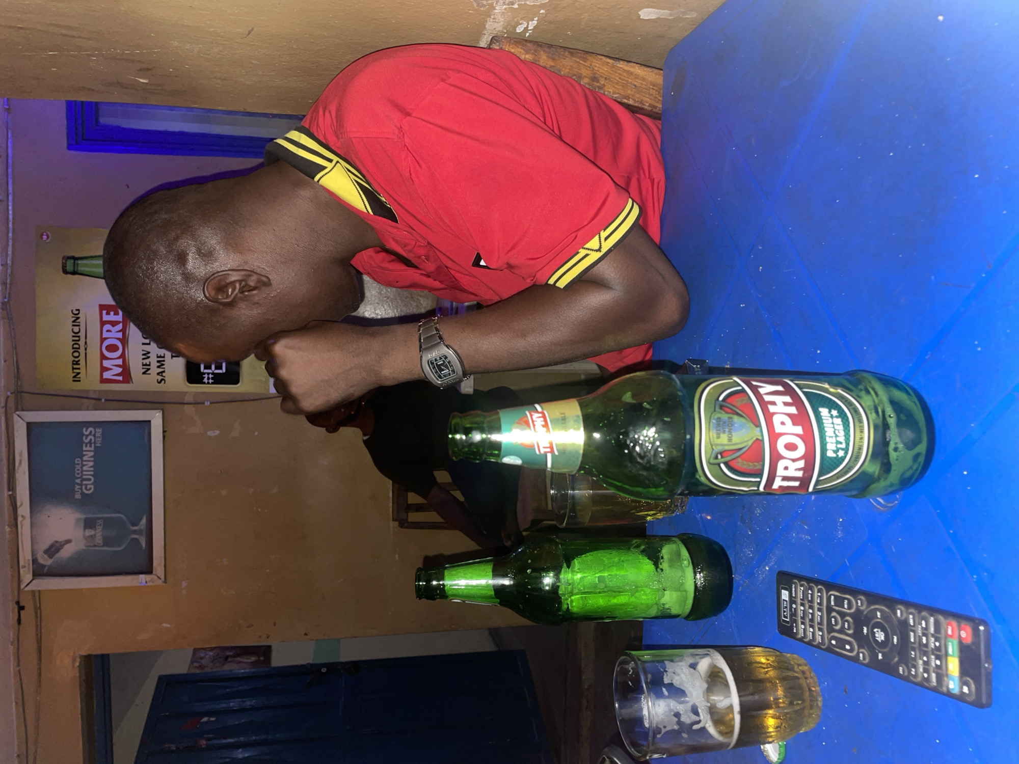 A man is seen sleeping at a table at an establishment in Jos, Nigeria. He is wearing a red shirt with yellow and black accents. On the blue table in front of him are a Trophy beer bottle, an empty green beer bottle, a glass with residue, and a black remote control. Behind the man, there is a Guinness poster on the wall, and another poster featuring the word "MORE". Another person is partially visible sitting behind him. The surroundings appear to be a dimly lit bar.