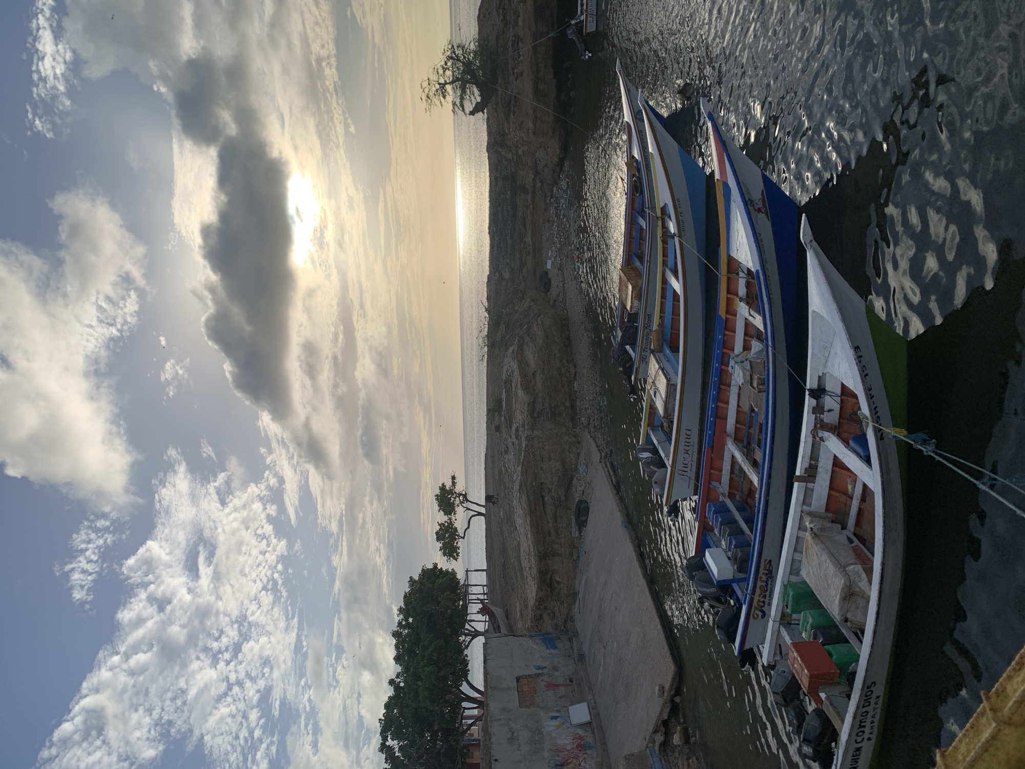 The image depicts a serene coastal scene in Juan Griego, Venezuela, with a focus on several small fishing boats docked in calm waters. The sky is a mix of blue with scattered white and grey clouds, with the sun setting or rising, casting a warm glow on the horizon. The water reflects the sky and boats, creating a textured surface.
In the foreground, a line of colorful fishing boats, with their outboard motors, are tied together. The boats are predominantly white with accents of blue, red, and orange along their hulls and interiors. Some boats contain equipment such as plastic containers and covered items. One boat in the front has text on its hull that reads "QUIEN COMO DIOS PAMPATAR" and "ARSH-PE 7673". Another boat is named "Josefina" and bears the text "PAMPATAR".
Behind the boats, a rocky shoreline leads to a small, dry beach area. Several trees, one notably gnarled and leaning, are scattered along the coast. A building with some graffiti on its exterior wall is visible on the left side of the image, adjacent to a concrete ramp leading into the water. The background shows the vast expanse of the sea meeting the horizon under the diffused sunlight. No people are clearly visible in the image. The overall impression is one of a quiet, working harbor at the end of the day.
