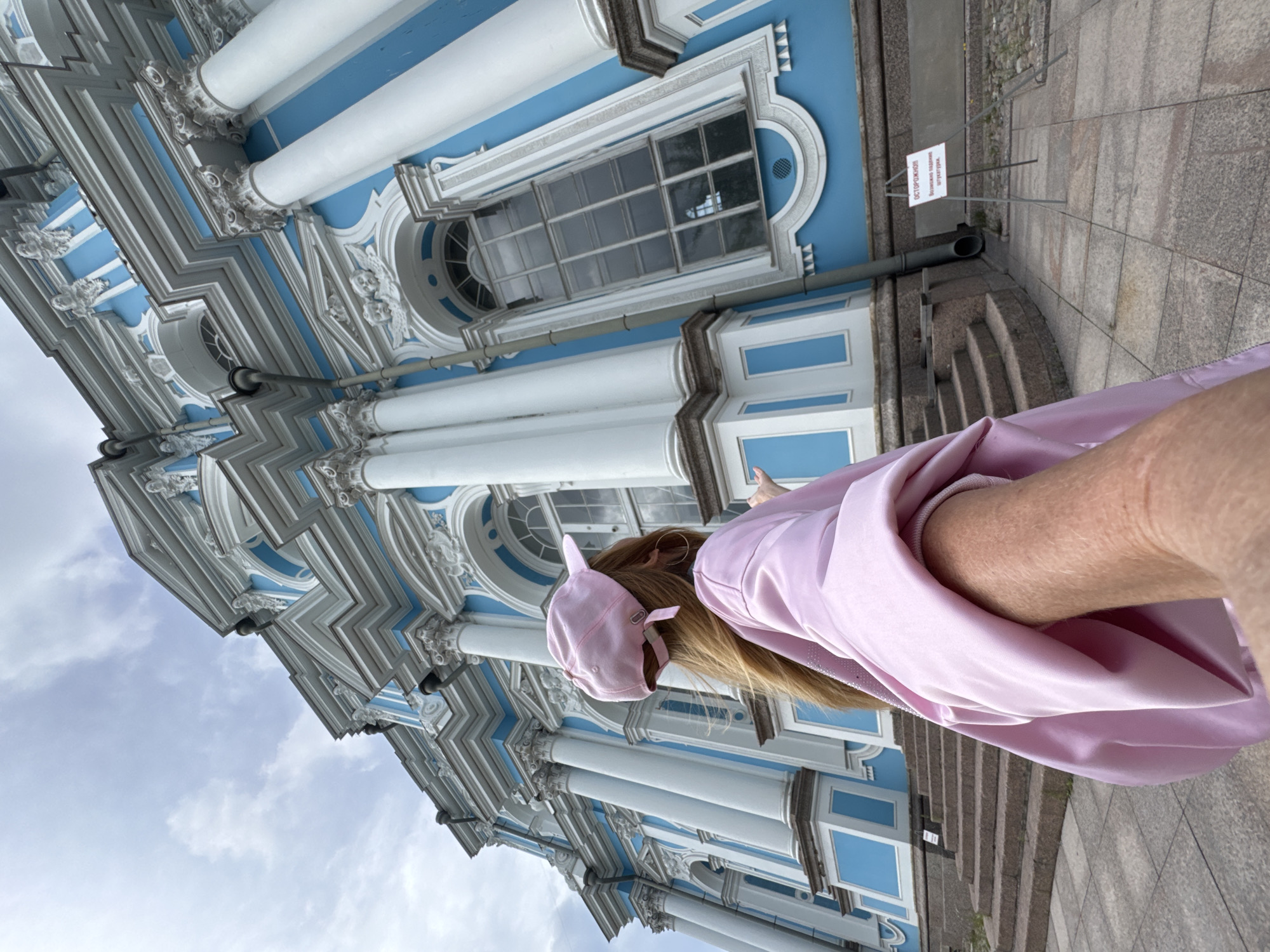 The image shows a person from behind, wearing a pink baseball cap with ear-like embellishments and a flowing pink outfit. They are standing in front of a grand blue and white building with ornate architectural details, including columns and intricate moldings. The building appears to be a historic structure, possibly a palace or a significant religious building.
The person is looking up at the building, with one hand outstretched slightly towards it. Their hair is long and blonde, and they are wearing a hoop earring. The overall impression is one of admiration or awe of the architecture.
The scene suggests it is daytime, with a cloudy sky. A sign with Russian text is visible on the ground to the right of the building. The ground is paved with large stone slabs, and there are stone steps leading up to the building. The person's posture and the way they are interacting with the environment suggest they are exploring or sightseeing.
The building's facade is a striking combination of bright blue and white, with a repeating pattern of columns and decorative elements. The windows are large and appear to be multi-paned. The architectural style hints at Baroque or Rococo influences, common in Saint Petersburg. The overall mood is serene and contemplative.
The visible text on the sign reads "ОСТОРОЖНО!" which translates to "CAUTION!". Below that, it says "Вход в здание ограничен." meaning "Entrance to the building is restricted."