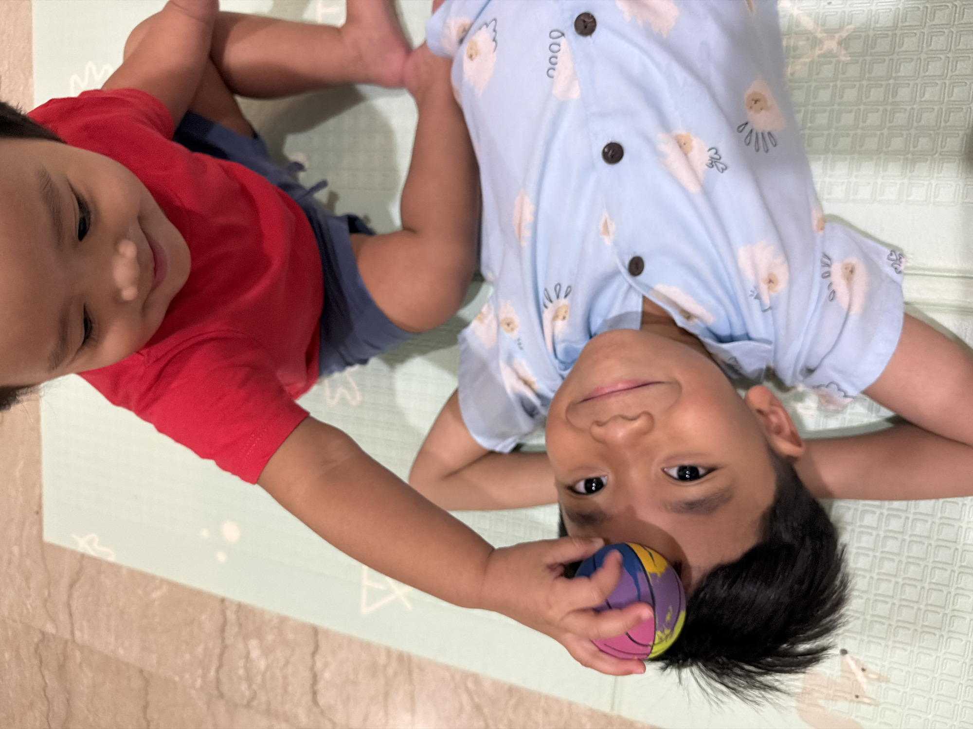 Two young children are on a light green mat with a patterned surface. The child in the foreground is lying on their back, wearing a light blue short-sleeved shirt with white sheep and clouds printed on it and two dark brown buttons. This child has dark hair and is looking up and to the left with a slight smile. The child's right arm is extended upwards, and they are holding a small, multicolored ball with their left hand. The child in the background is sitting up and leaning forward, wearing a red short-sleeved t-shirt and blue shorts. This child is reaching down towards the child on the mat, with their right hand near the ball. The background appears to be a floor with a marble-like pattern. The image does not provide any visual cues to determine the location in the city of Central Jakarta, Indonesia.