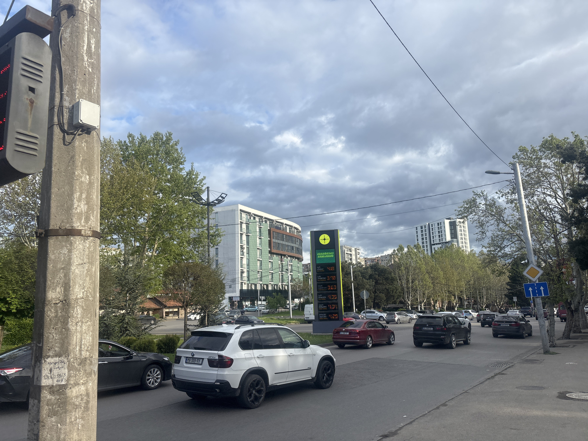 The image captures a street scene in Tbilisi, Georgia, showcasing vehicular traffic and urban infrastructure. A white BMW X5 and other cars travel along the road, adjacent to a petrol station displaying fuel prices. The architecture in the background includes modern buildings with green accents. A traffic light and utility pole are visible on the left. Trees line the street, adding greenery to the environment under a cloudy sky.