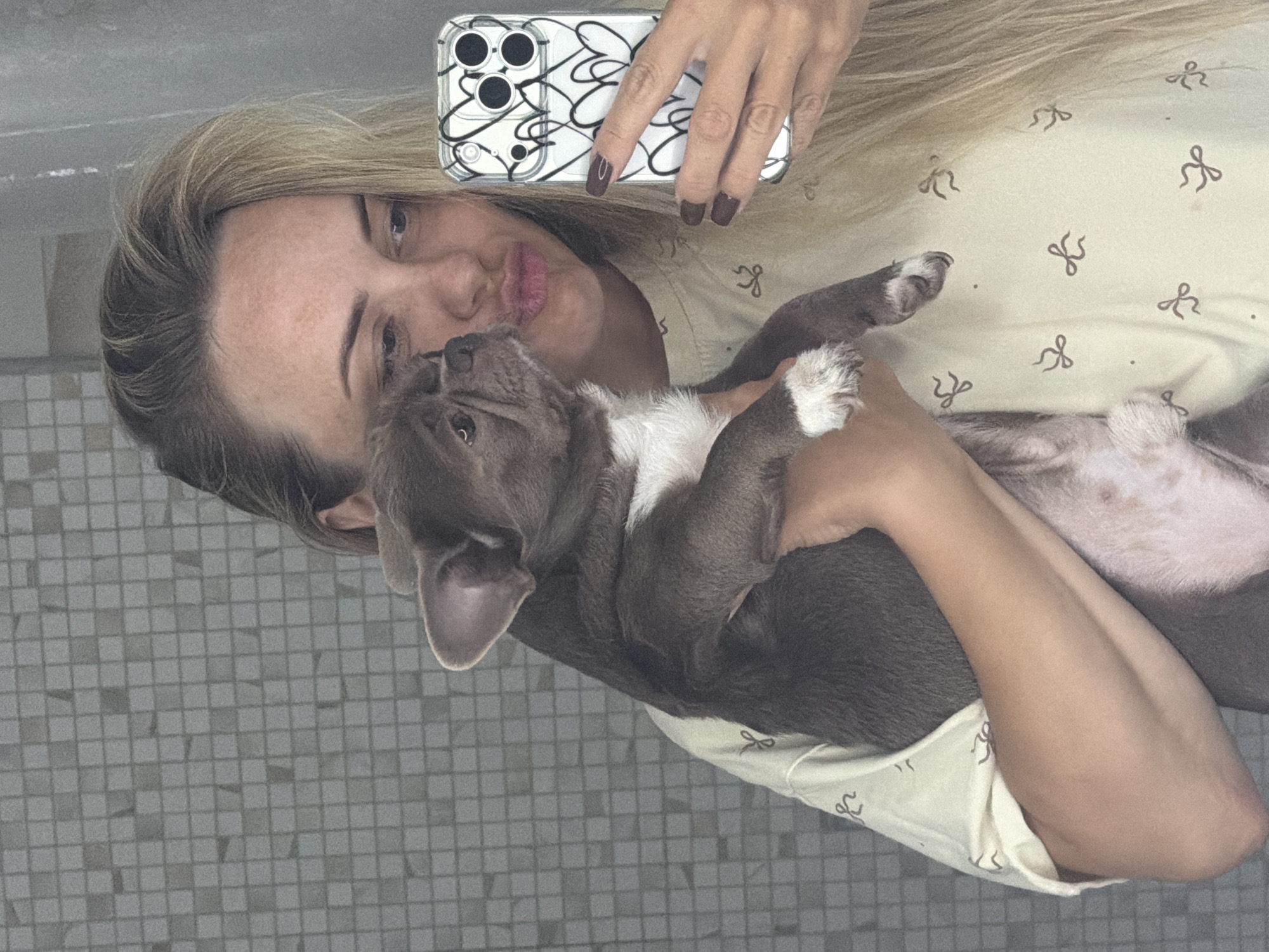 This media file is a selfie taken by a woman holding a puppy. The woman has blonde hair and is looking at the camera with her lips puckered. She is wearing a light-colored shirt with a bow pattern. The puppy is gray and white with floppy ears. It is looking up at the woman. The background appears to be a tiled wall, possibly in a bathroom or kitchen. The image is taken from a slightly overhead angle, and the lighting suggests it might be daytime. The overall mood seems affectionate and playful. No visible text is present in the image. The location context provided is Votkinsk, Russia.
