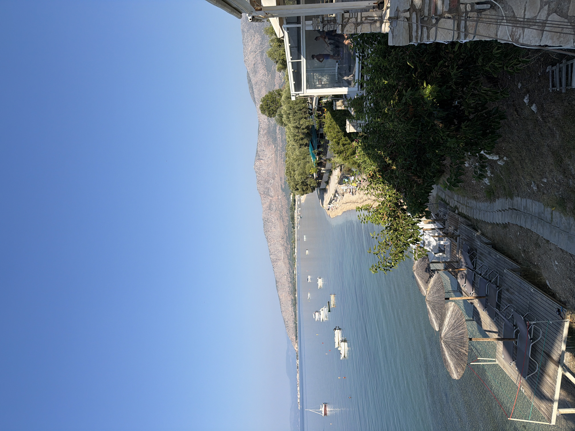 The image depicts a scenic view of the Municipal Unit of Alyzia, Greece, on a clear, sunny day. The foreground features a wooden deck with several lounge chairs and thatched umbrellas set up for relaxation. The deck extends over the calm, turquoise water, with a gently sloping sandy beach visible to the left.
In the middle ground, the sea is dotted with several small white boats and one sailboat with a red sail. A lush green coastline runs parallel to the water, lined with trees and vegetation, and a few buildings are nestled among the greenery, some with awnings providing shade.
The background is dominated by a range of rugged, mountainous terrain with sparse vegetation, stretching across the horizon under a clear blue sky. On the right side of the image, part of a building with a stone facade and glass windows is visible. Inside the building, two individuals are present, one appearing to be in athletic attire. The overall scene suggests a tranquil seaside resort or a coastal village setting in Greece.